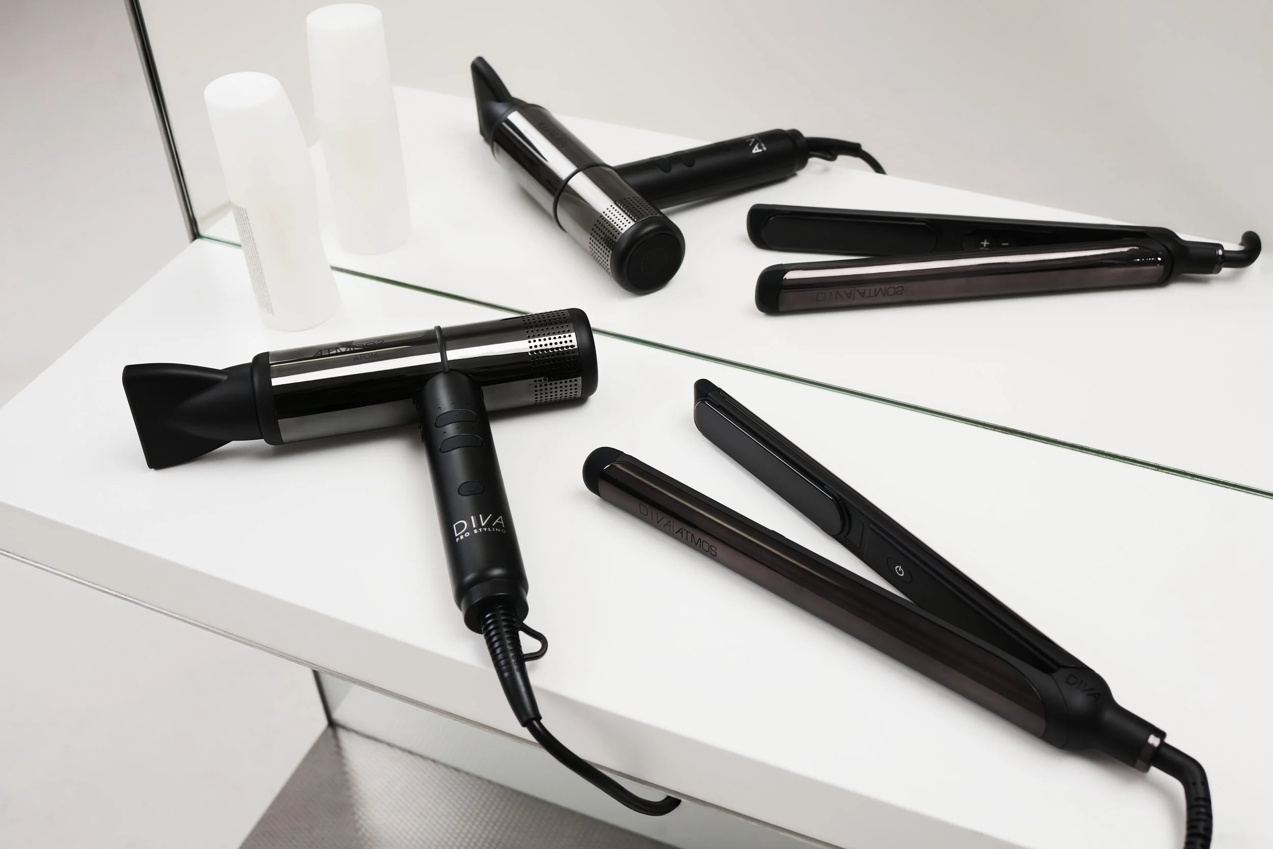 Various hair styling tools including a blow dryer, flat iron, curling iron, and hot air brush on a white table with a mirror in the background.