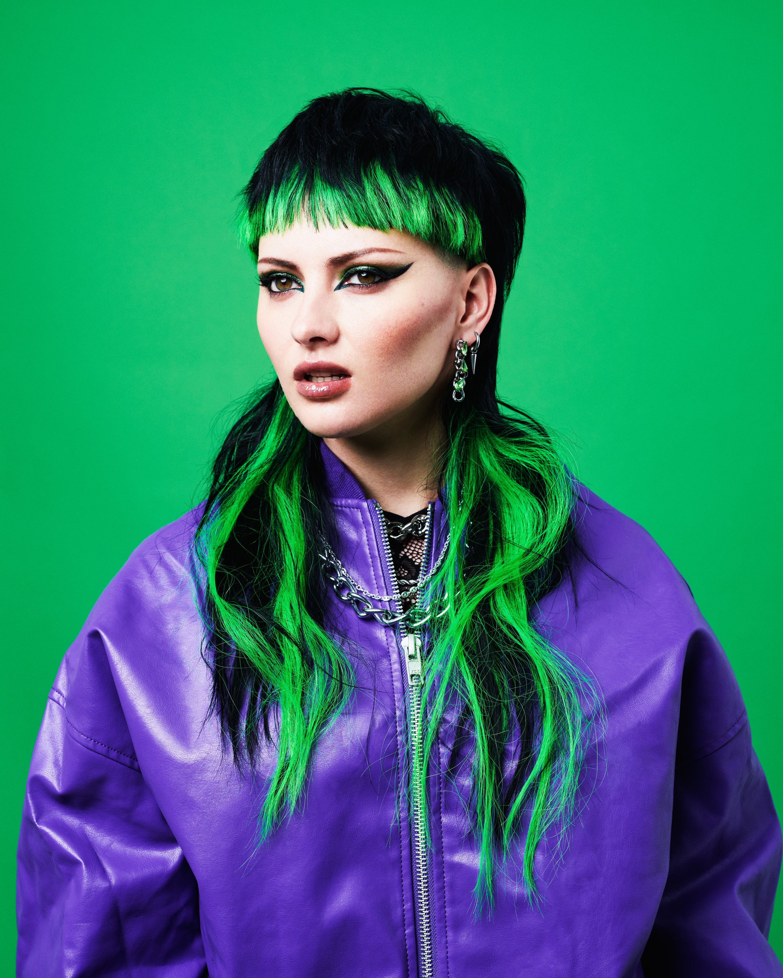 A woman with black and green hair, wearing dark eye makeup, large earrings, a silver chain necklace, and a purple jacket, standing against a green background.