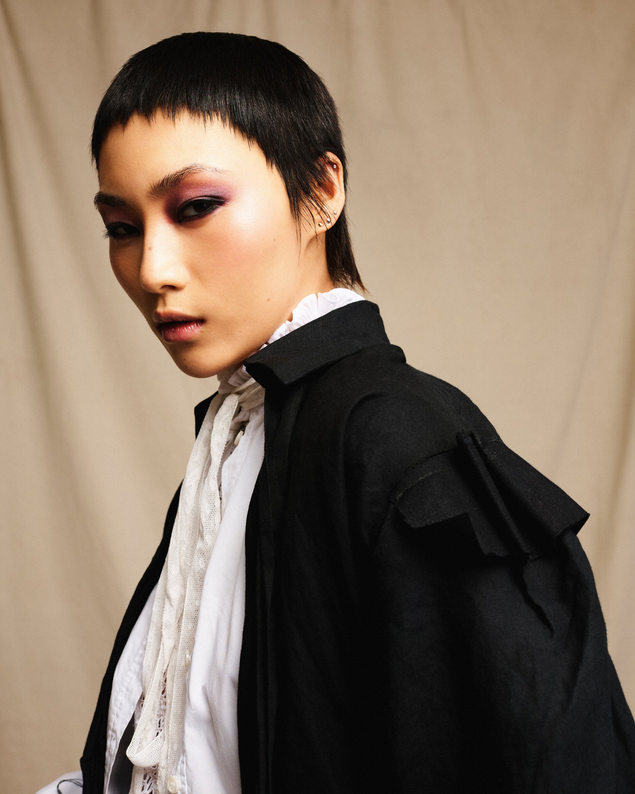 A young woman with short black hair and earrings, dressed in a black blazer and white blouse with lace details, posing against a neutral background.