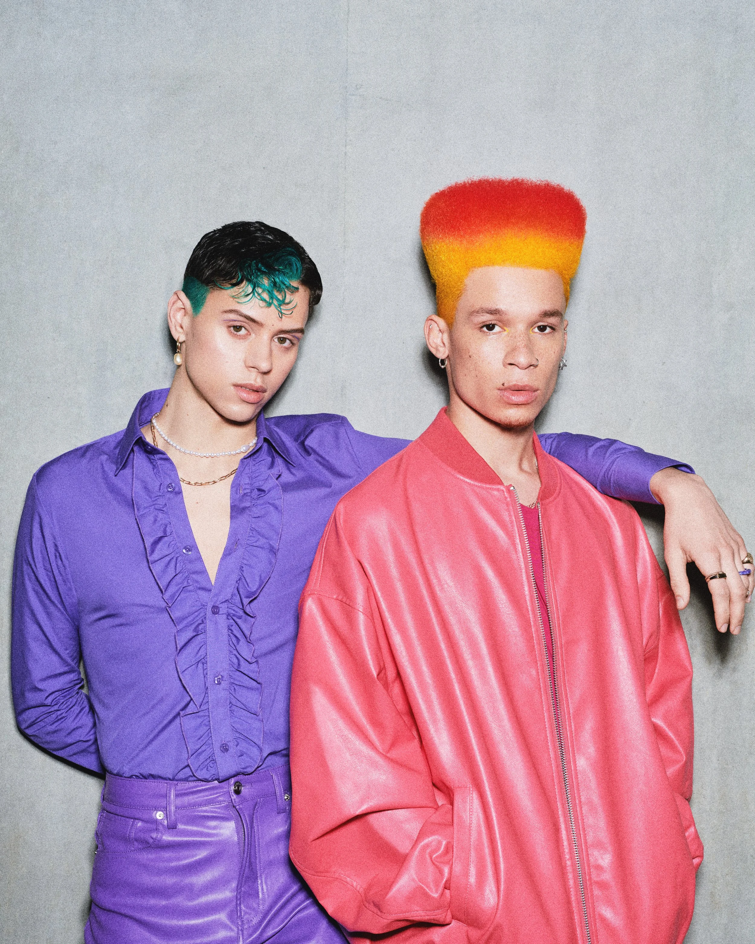 Two individuals with colorful hair and clothing posing against a plain background.