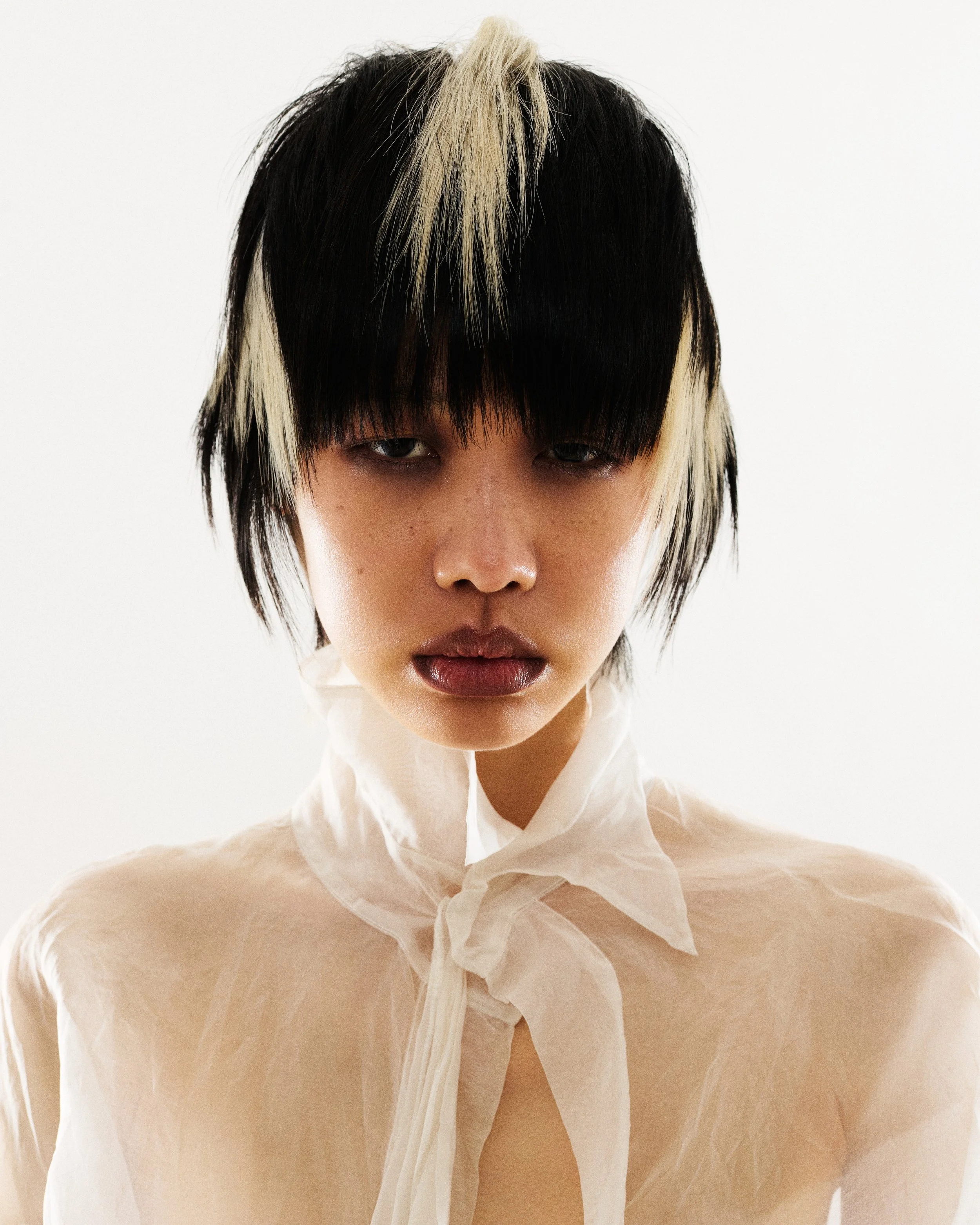 Close-up of a person with short black hair with blonde highlights, wearing a sheer white fabric draped around their neck and shoulders, against a plain white background.