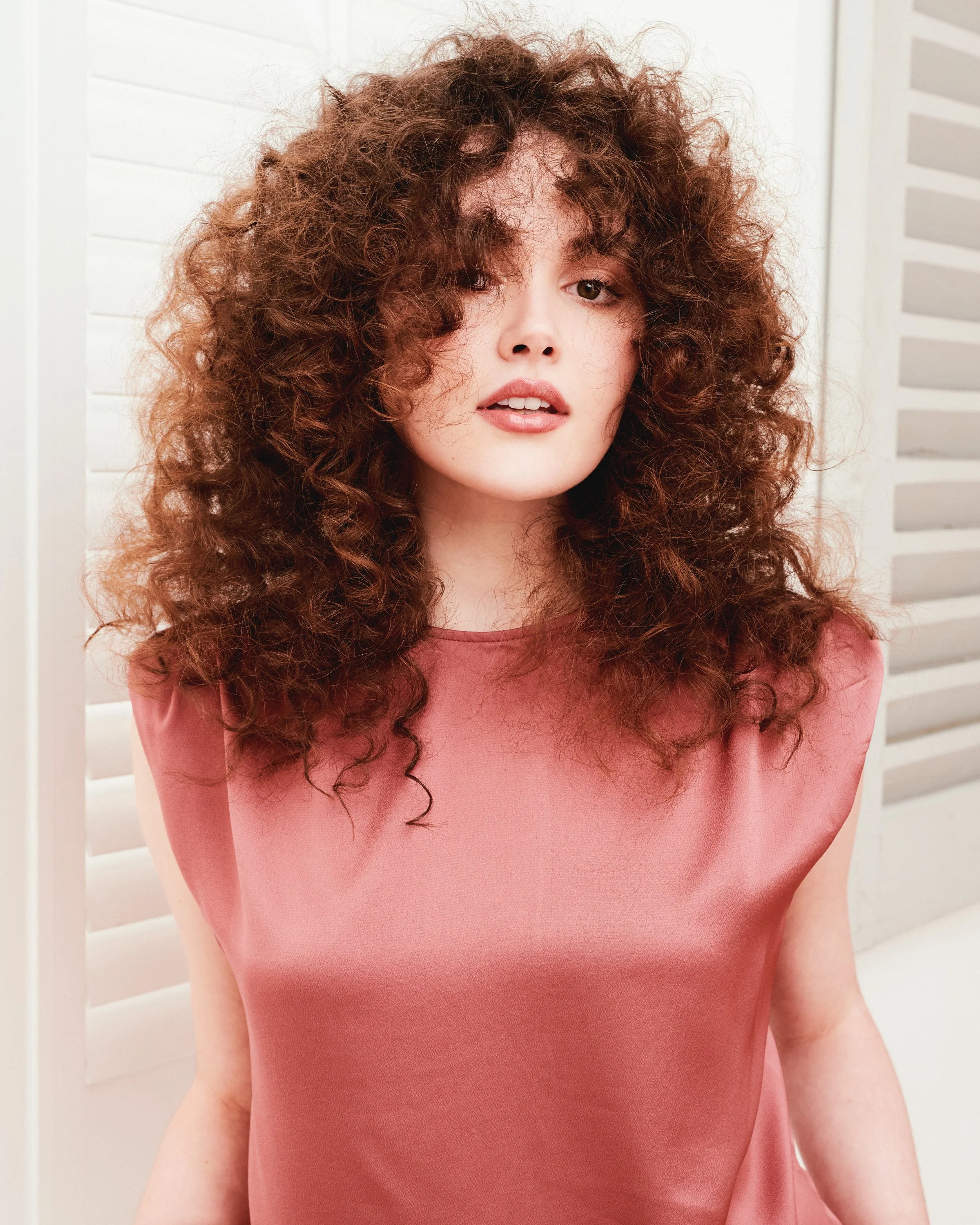 A woman with curly brown hair wearing a pink top standing in front of a white background with horizontal blinds.