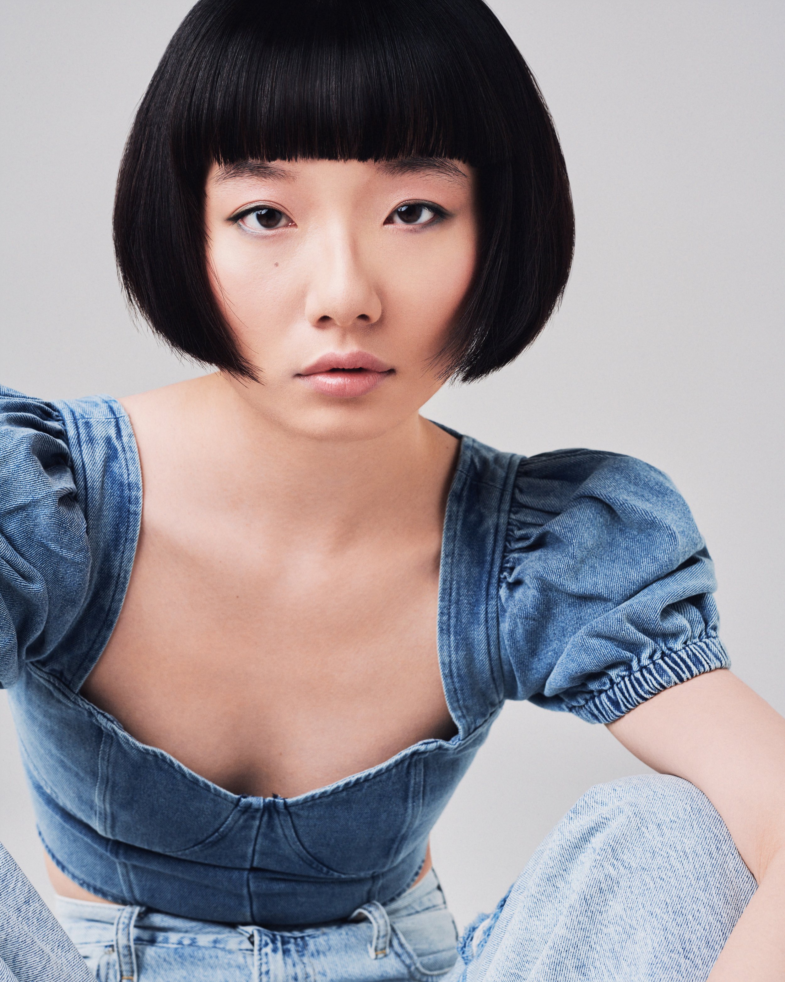 A young woman with short black hair and a bowl haircut poses with her head tilted slightly, wearing a denim shirt and jeans against a plain background.