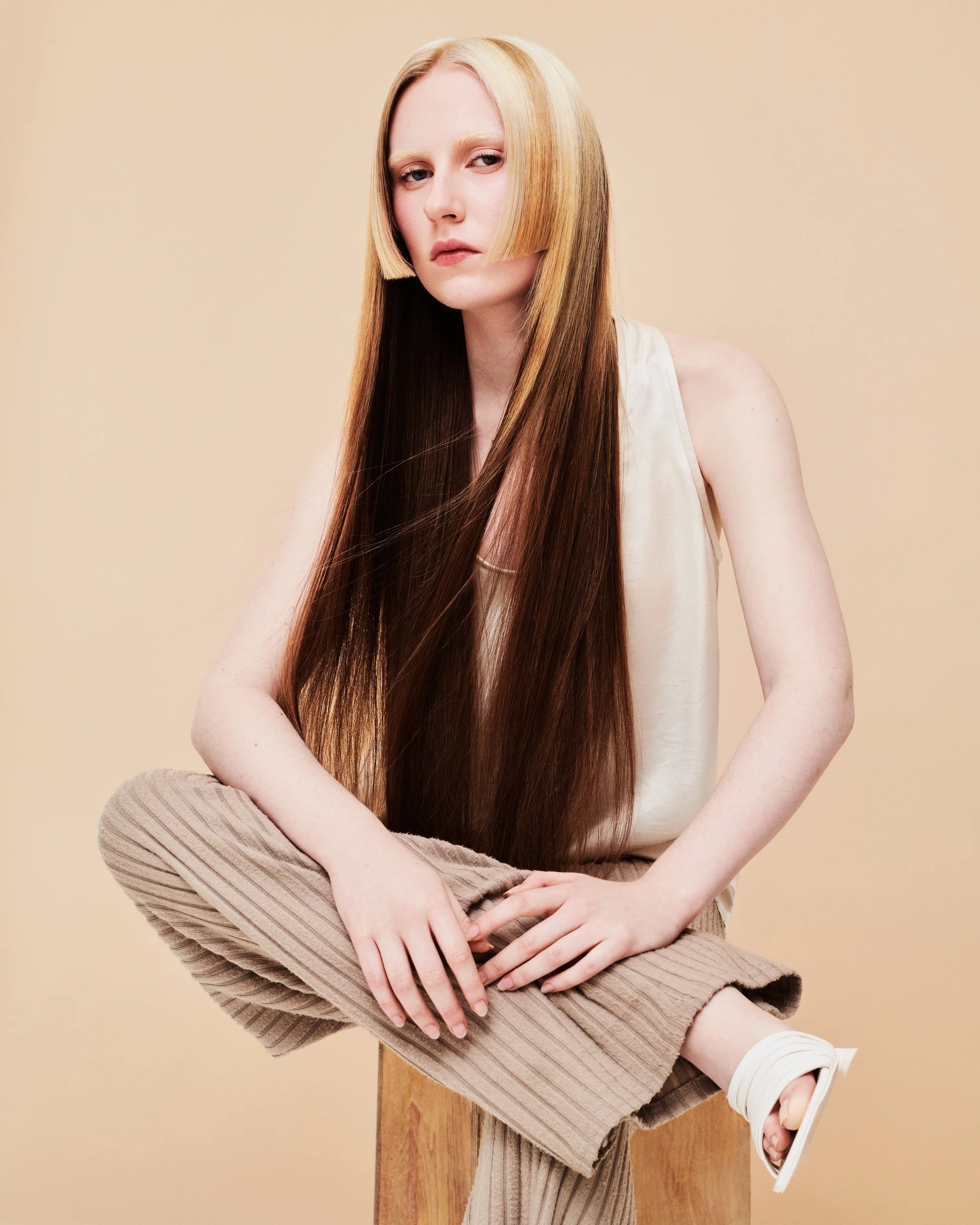 Young woman with long straight hair, sitting on a small wooden stool, wearing a beige sleeveless top and brown striped pants, with a neutral expression, against a beige background.