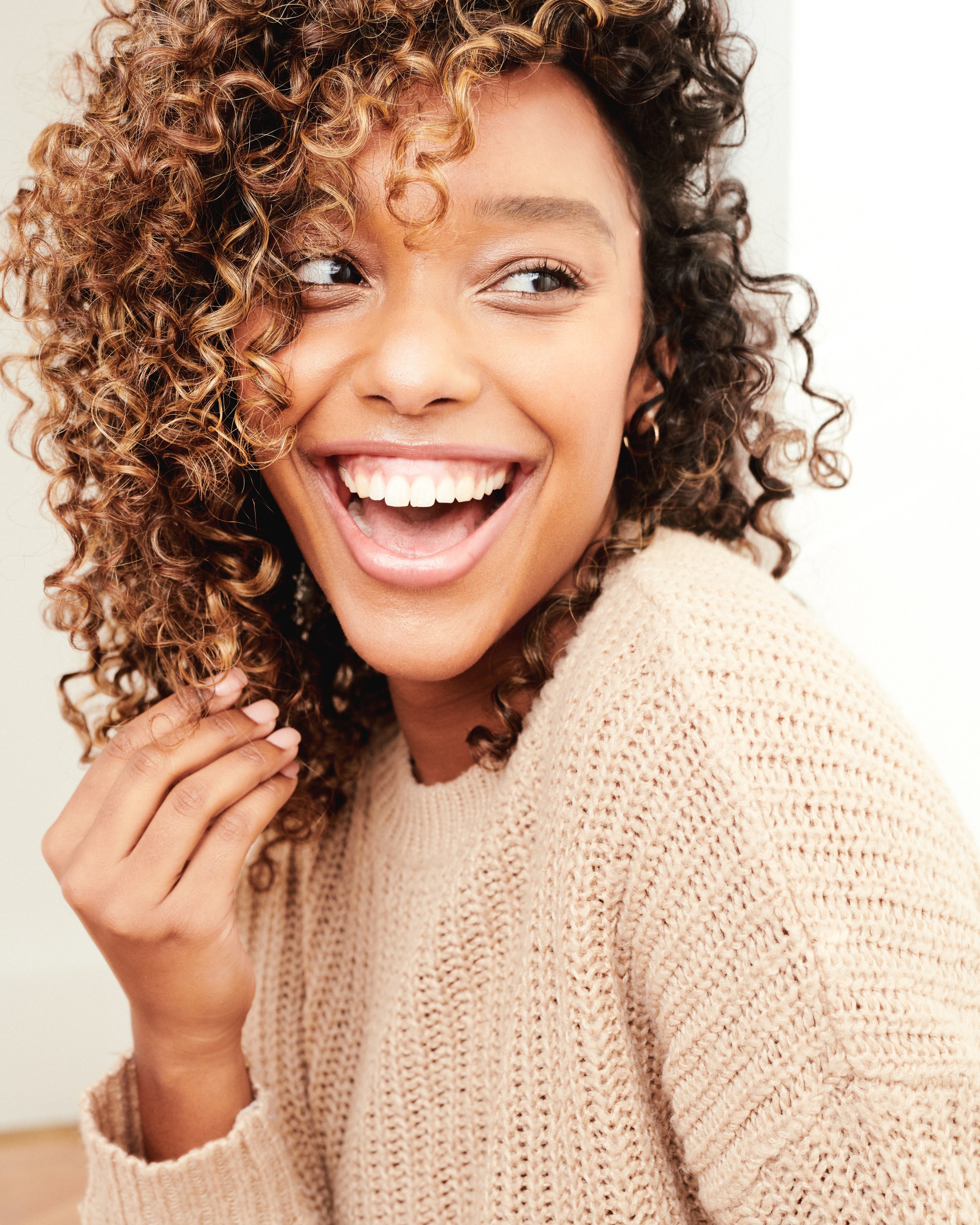 A woman with curly hair smiling and laughing, wearing a beige knit sweater, with her hand touching her shoulder, in a bright indoor setting.