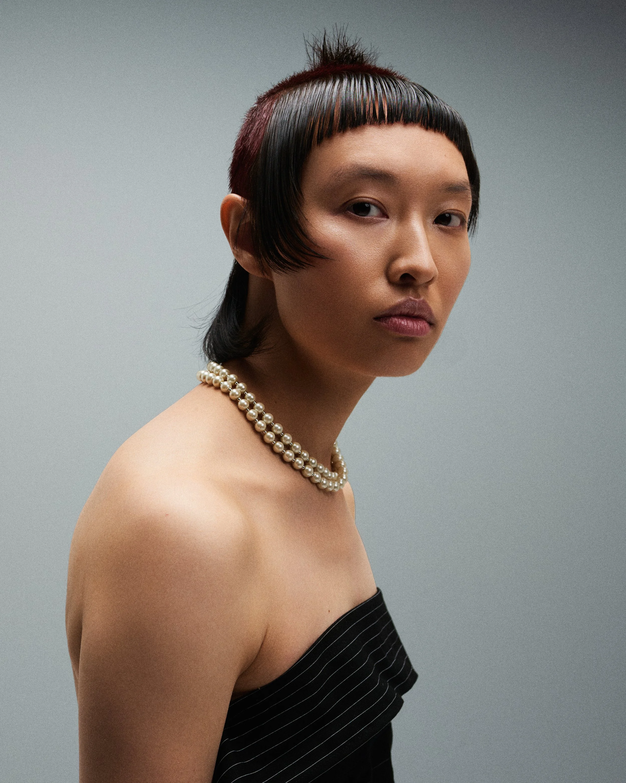 Close-up of a woman with short black hair and blunt bangs, wearing a pearl necklace, a black strapless dress with white pinstripes, and minimal makeup, against a plain gray background.