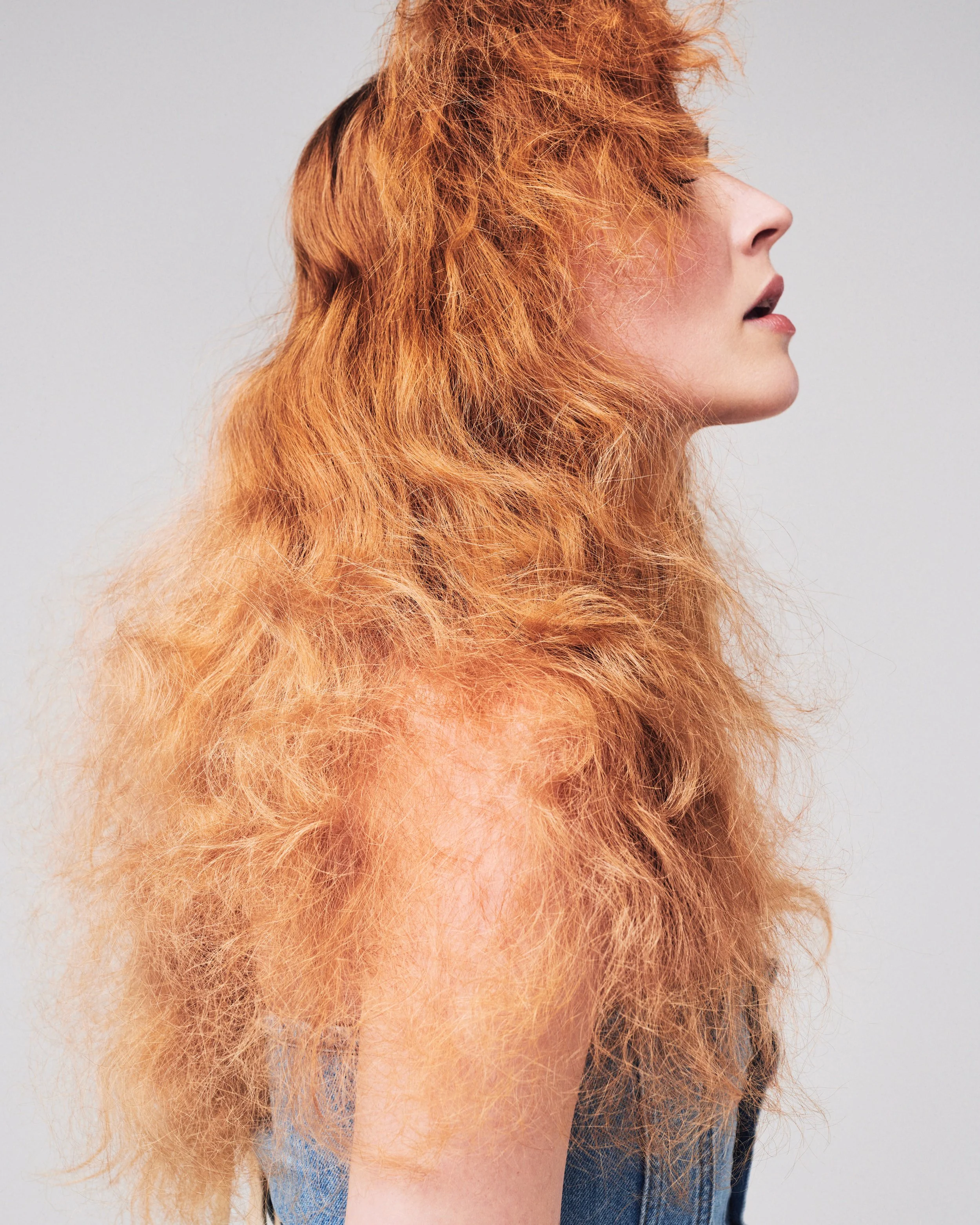 Side profile of a woman with bright orange, voluminous, curly hair, wearing a denim jacket against a plain light background.