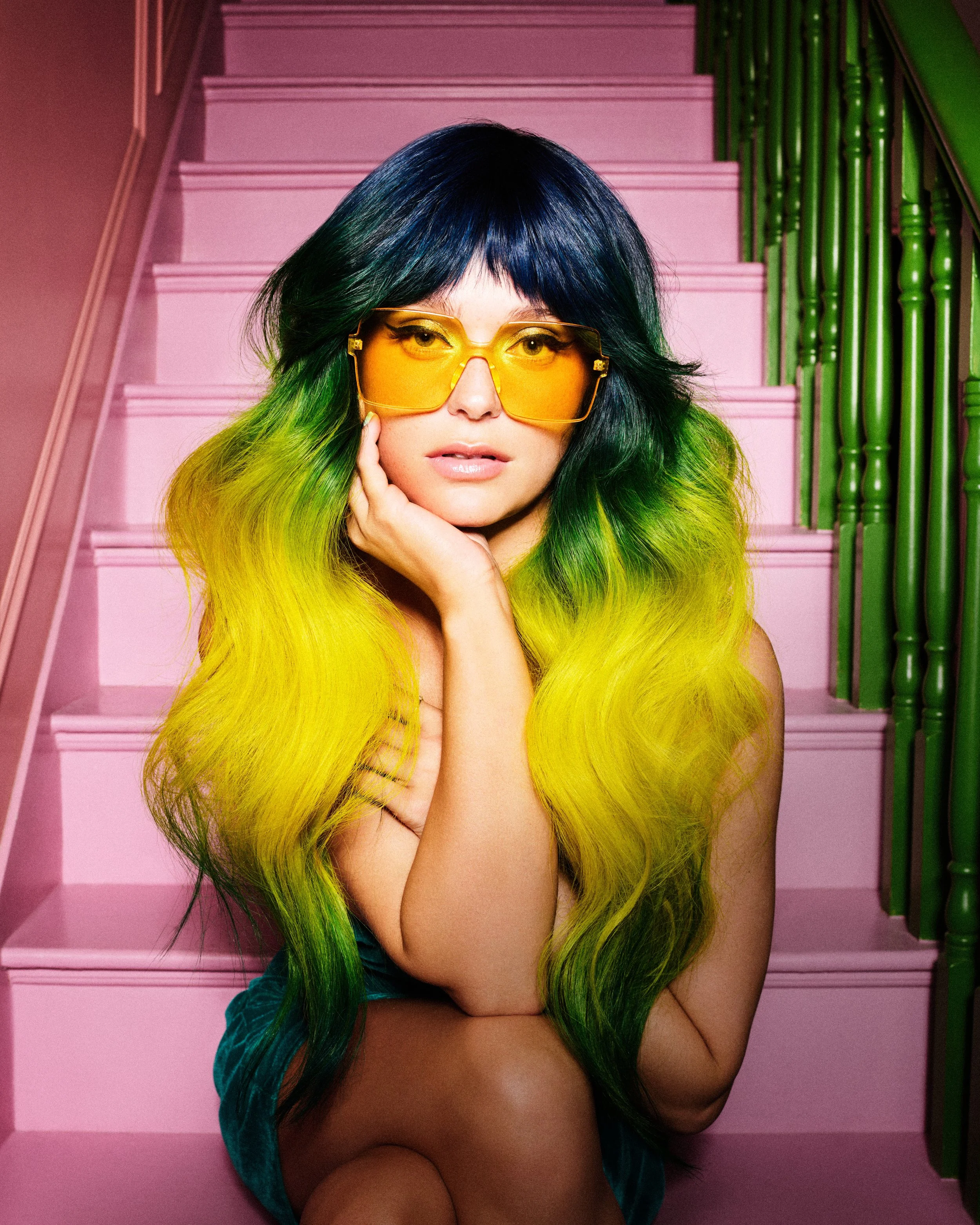 A woman with long, wavy hair that is black at the top and transitions to yellow and green towards the bottom, wearing oversized yellow-tinted sunglasses, sitting on pink stairs with green railings in the background, resting her chin on her hand.