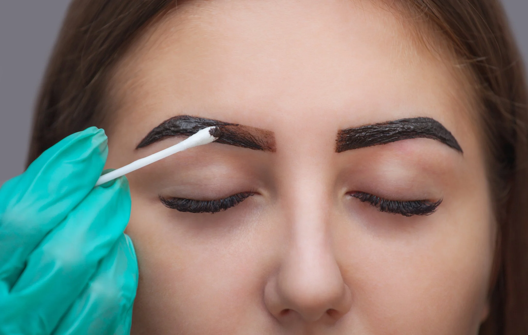 A woman with her eyes closed getting her eyebrows tattooed by a professional, using a cotton swab and gloves.