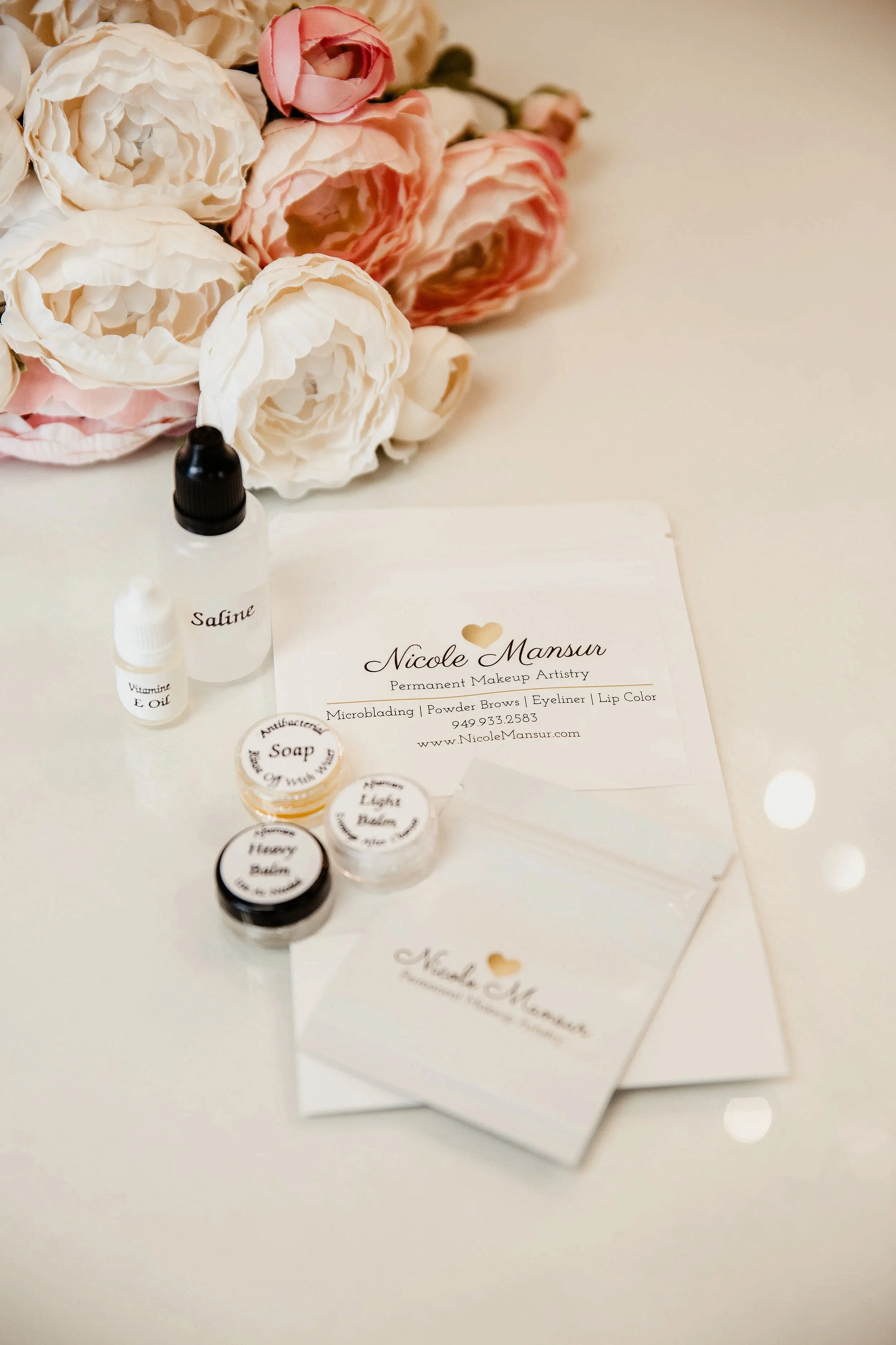 Business cards and supplies for Nicole Mansur, a permanent makeup artist, along with small containers labeled for different makeup pigments, a bottle labeled 'Saline,' a small bottle labeled 'Vitamin E Oil,' and a bundle of pink and white flowers on a white surface.