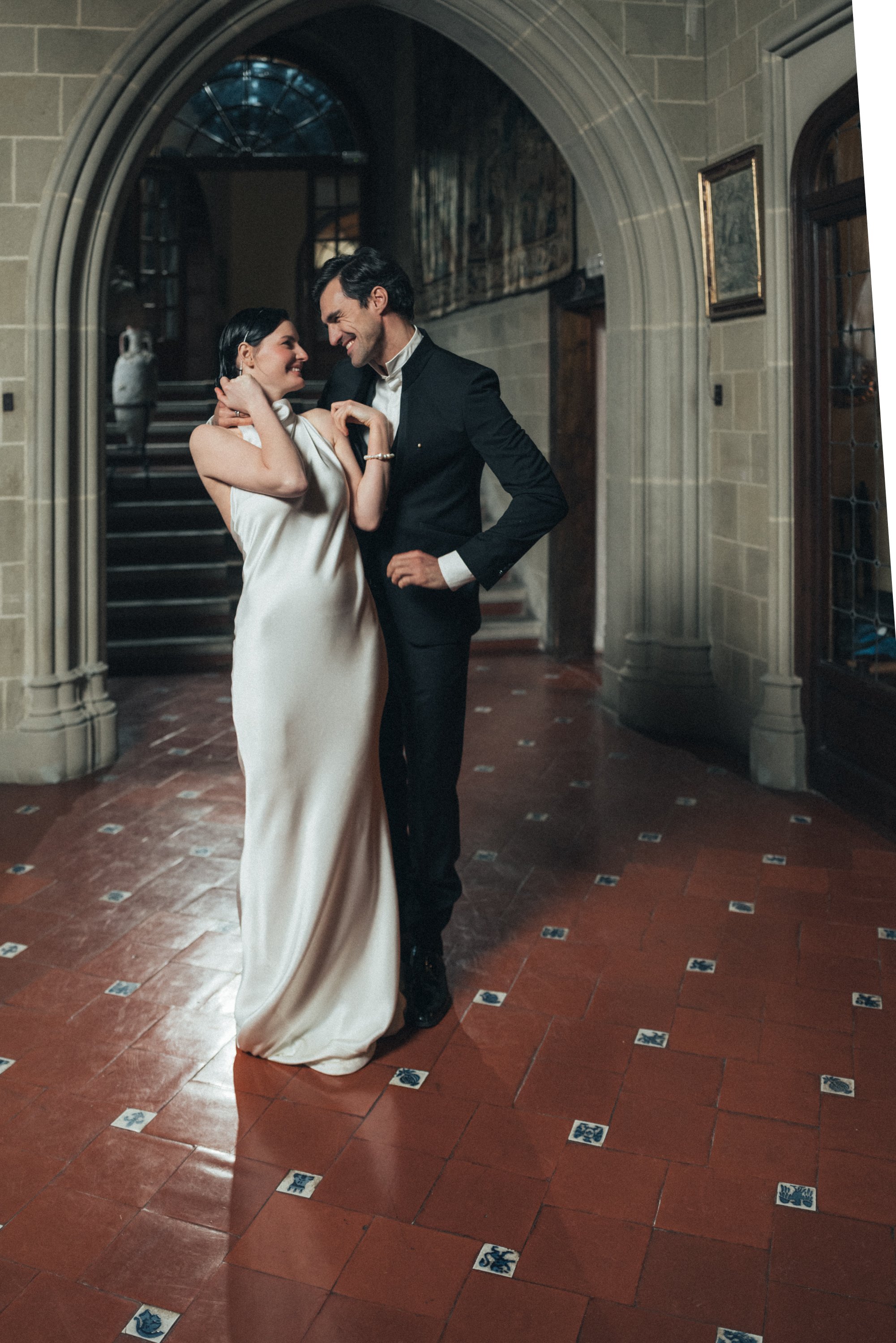 Elegant couple dancing in a formal setting, with woman in a white dress and man in a black suit, under a stone archway with tiled floor.
