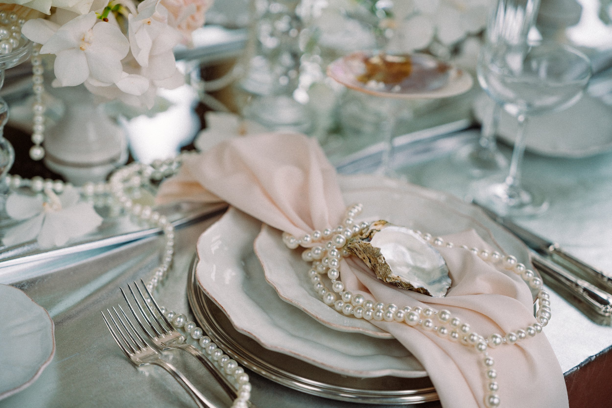 Elegant table setting with white plates, pearl necklace, pale pink napkin, and white flowers on a mirrored surface.