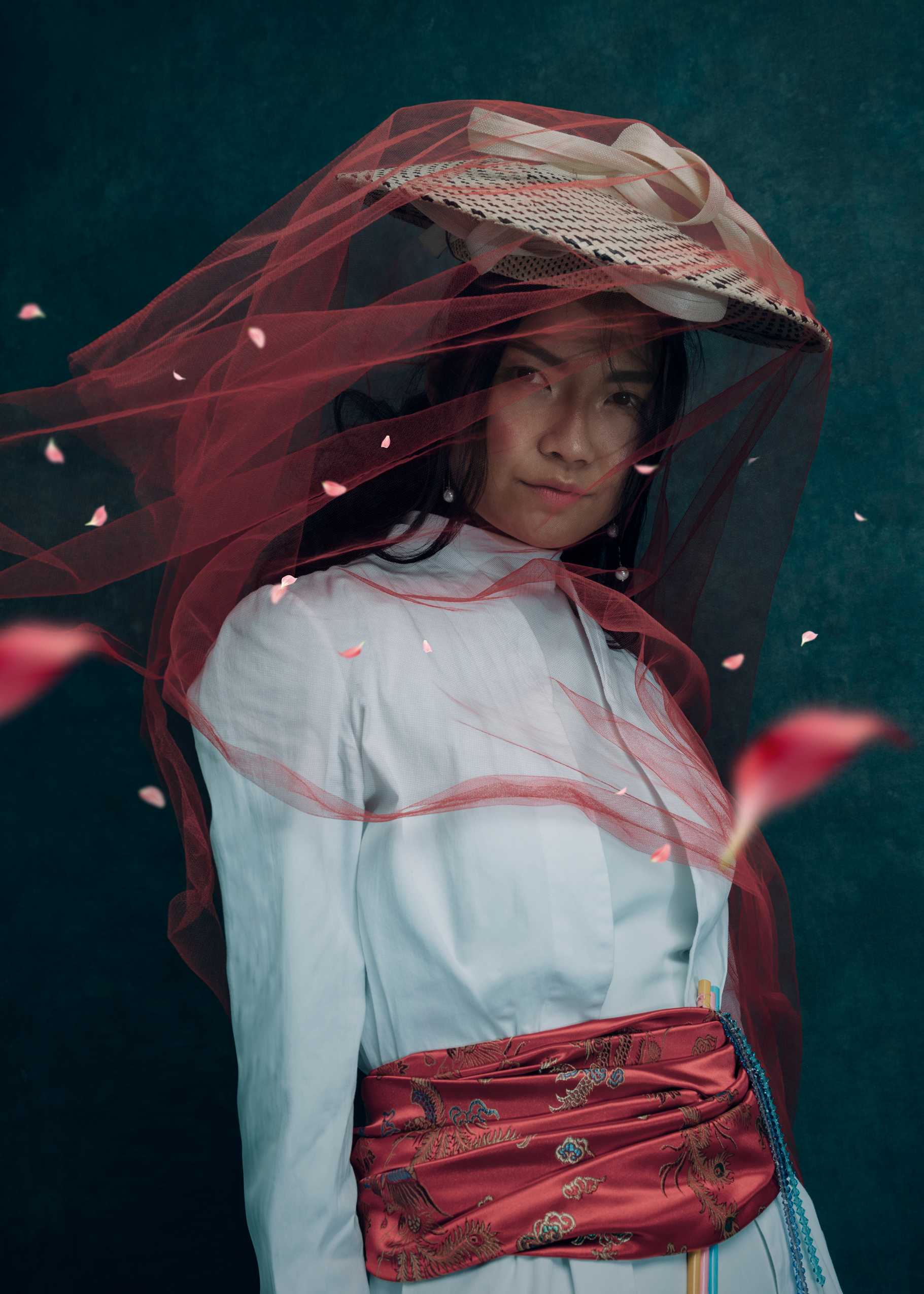 Person in traditional attire with red veil and straw hat, wearing a white top and red decorative sash, surrounded by floating petals.
