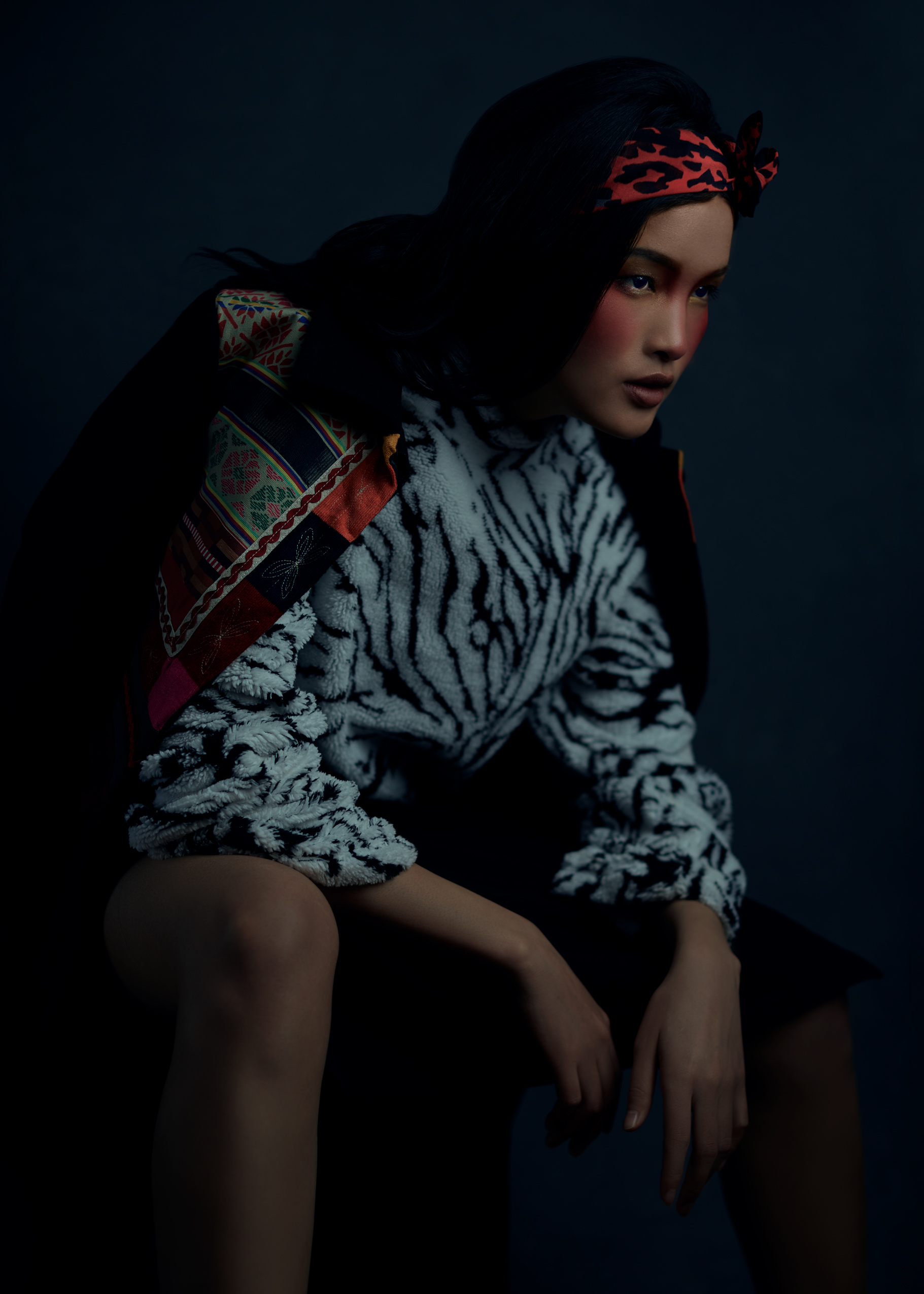 Fashion model sitting wearing zebra-patterned sweater, dark coat, and leopard-print headband, against a dark background.