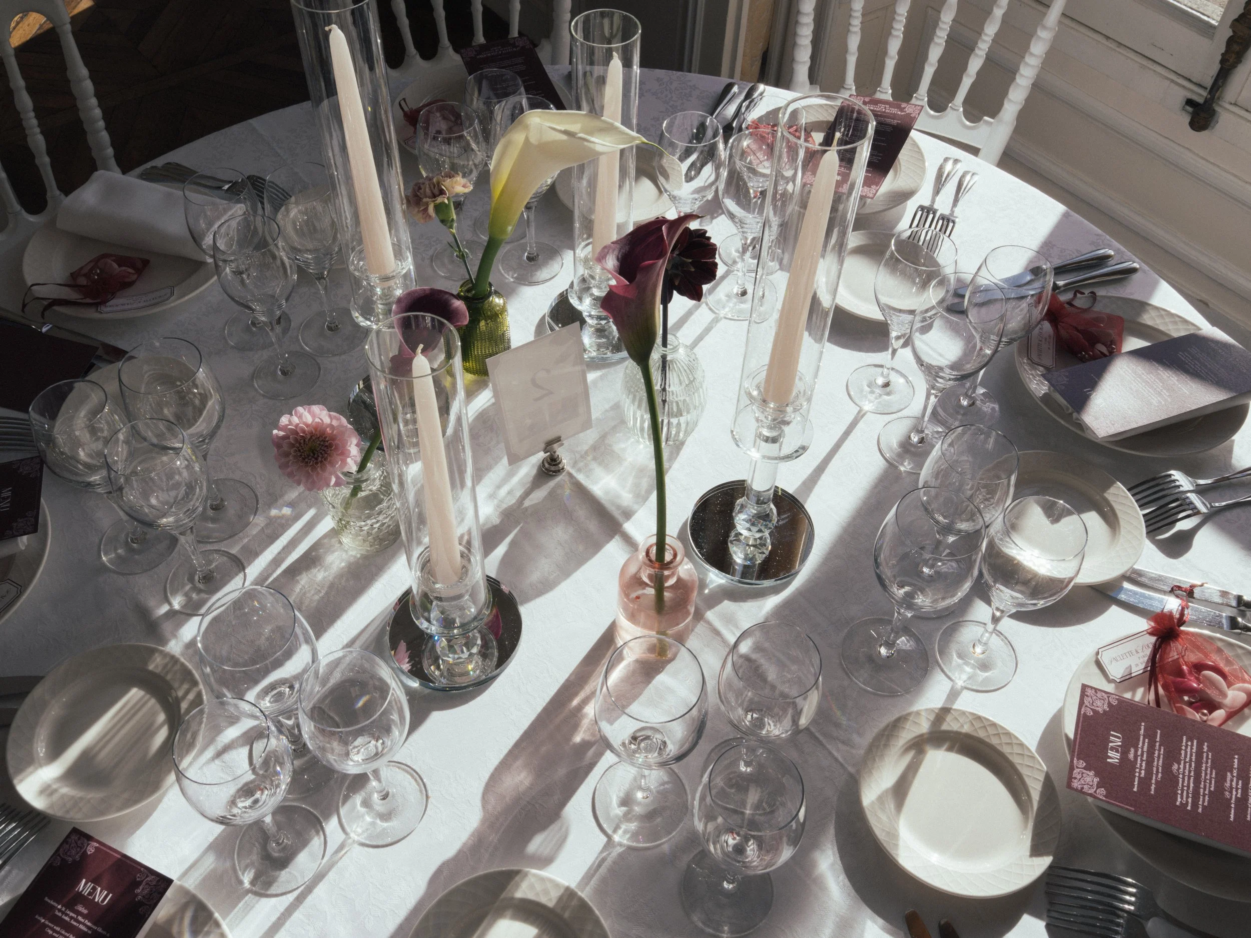 Round dining table set with white tablecloth, multiple clear wine glasses, white plates, napkins, silver utensils, and a central floral decoration with calla lilies and other flowers in glass vases, illuminated by sunlight.