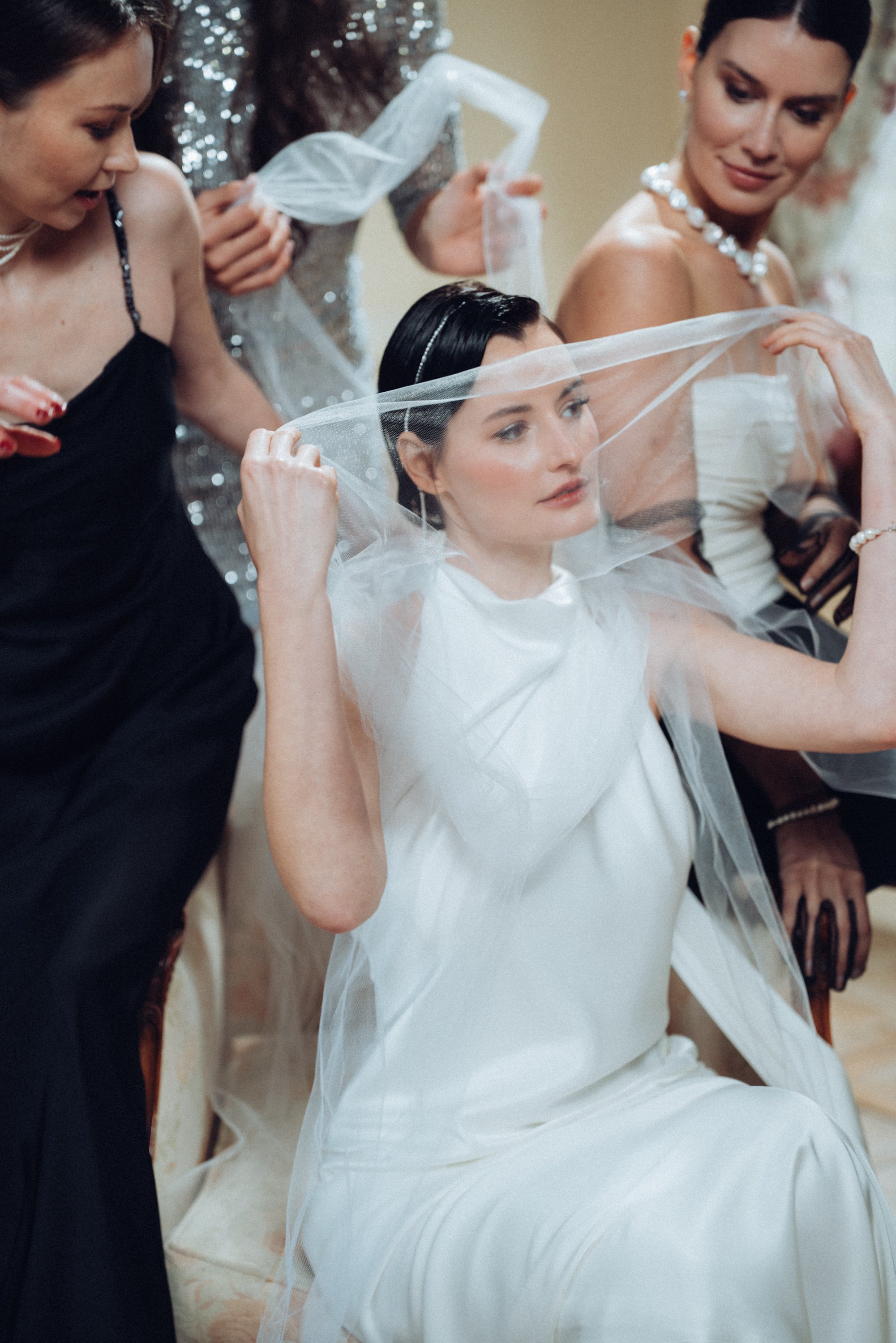 A bride in a white dress adjusting her veil while seated, surrounded by women in formal attire.