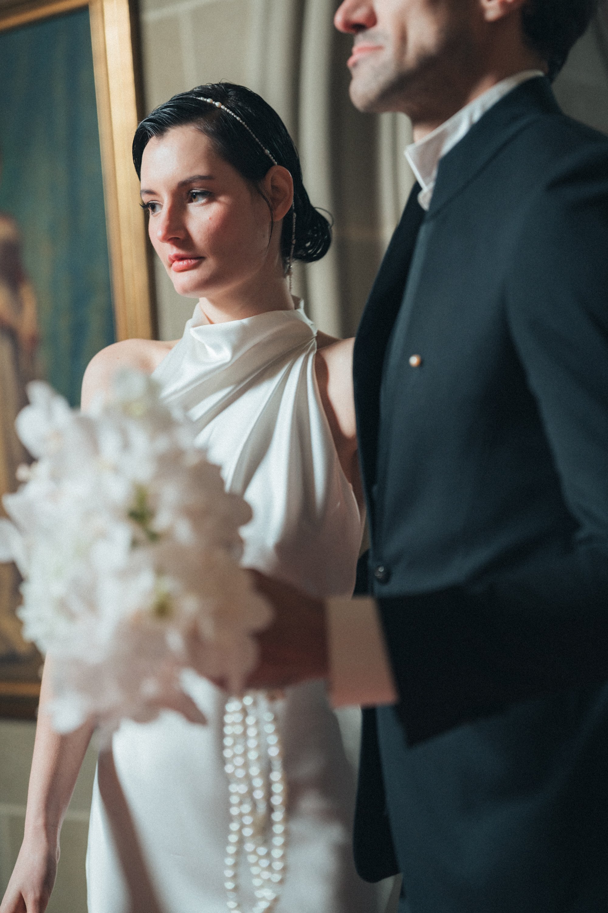A bride in a white dress holds a bouquet with ribbons beside a groom in a dark suit, standing in front of a framed painting.