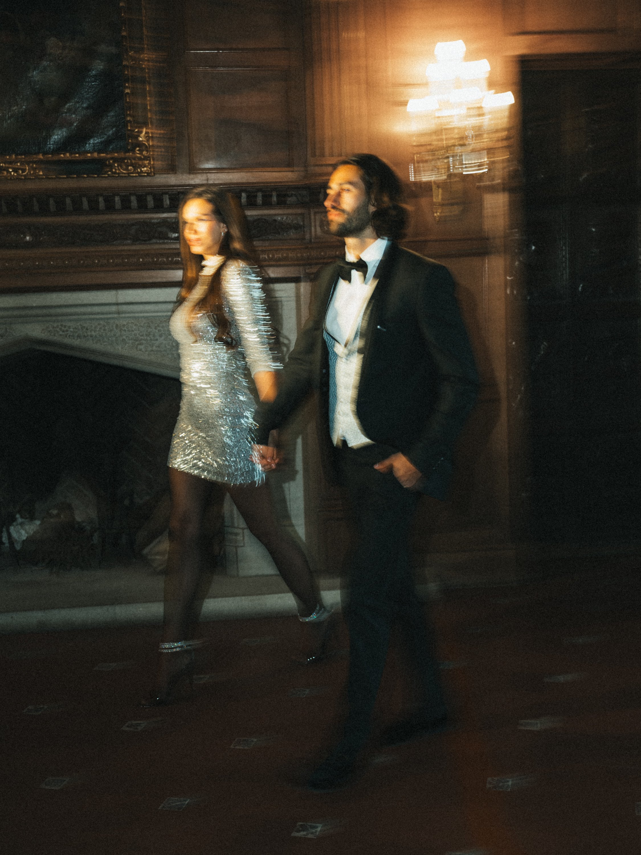 A man in a tuxedo and a woman in a silver dress walking in a dimly lit room.