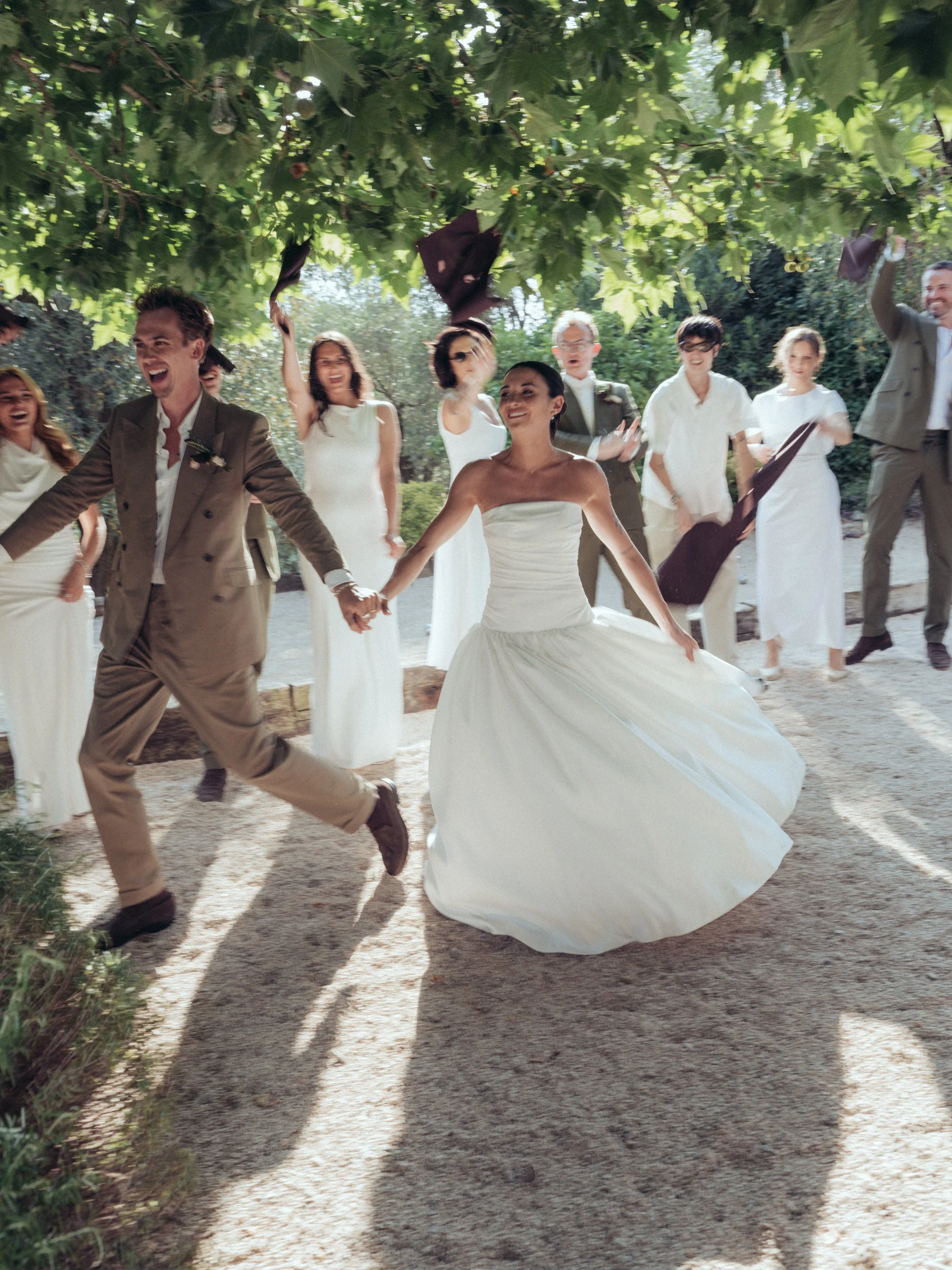 A bride and groom holding hands and dancing outdoors, surrounded by friends and family, with some tossing their hats in the air, in a lush green setting. Beautiful moment at their wedding in Provence at the french riviera.