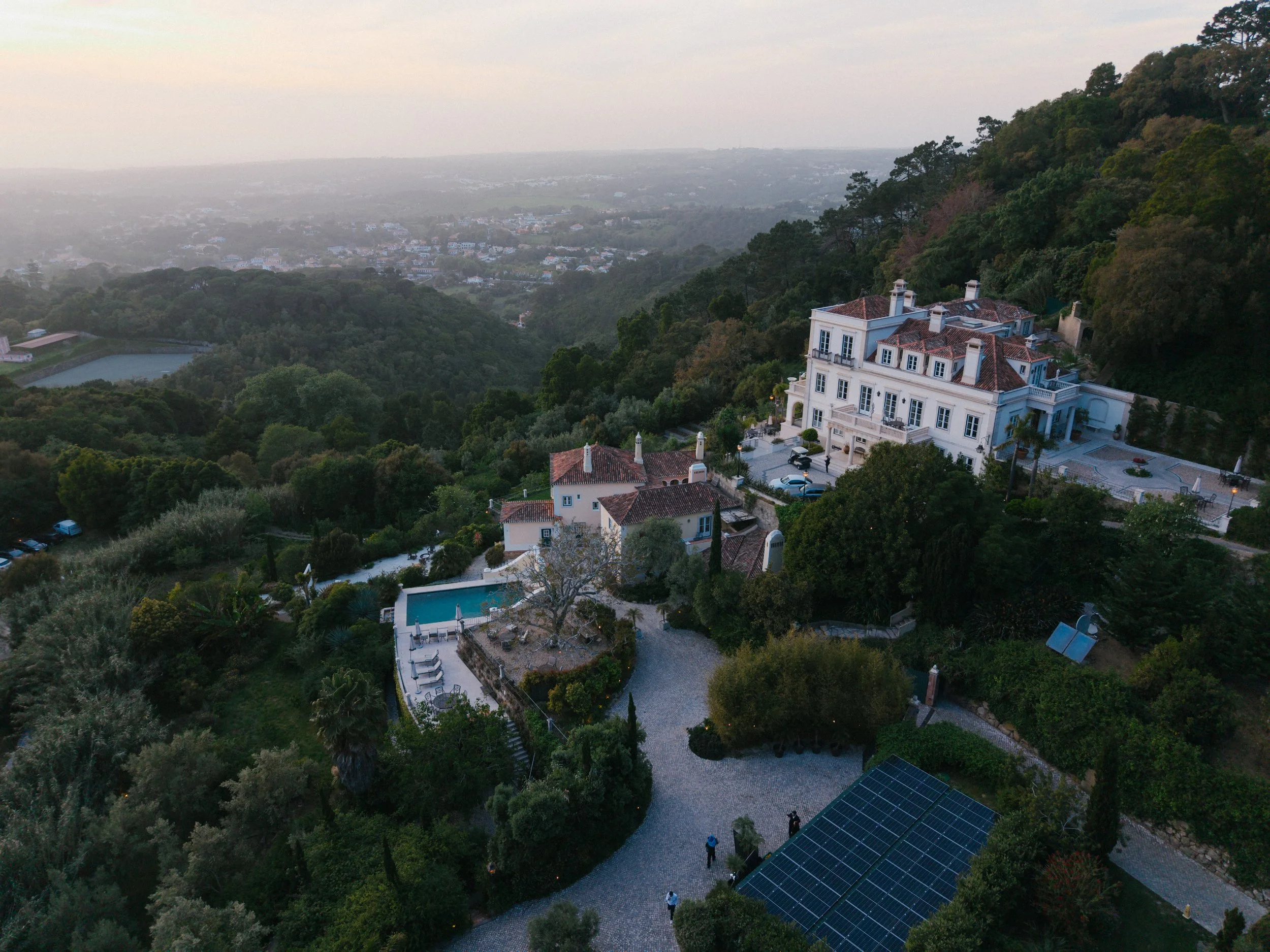 A large white mansion with red-tiled roofs and multiple chimneys sits on a hillside surrounded by lush green trees. There is a courtyard with a circular gravel driveway, several outdoor seating areas, and solar panels on a lower roof. The scene is set during dusk with a view over a valley filled with trees and distant houses, with a hazy sky.