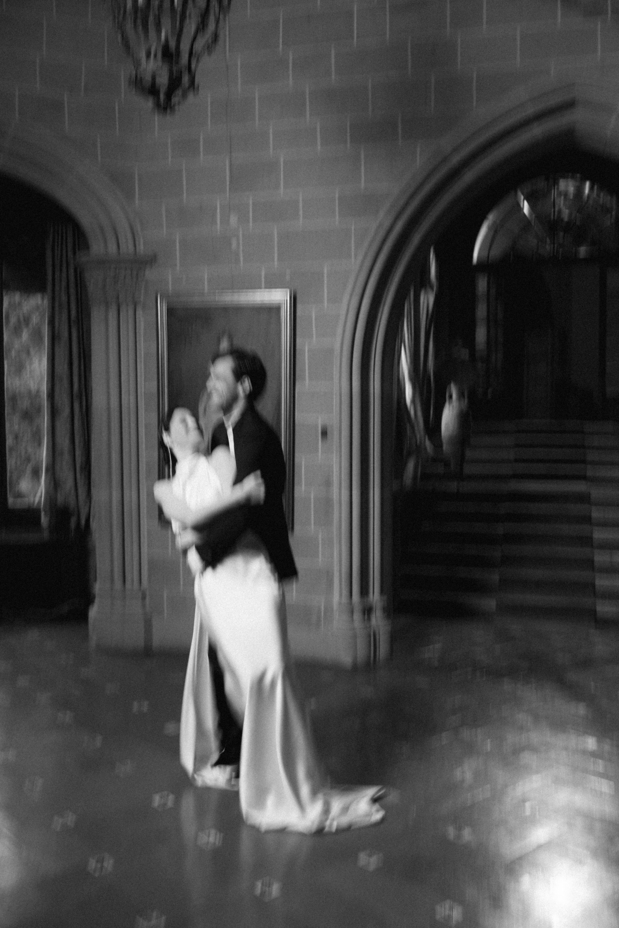 Black and white image of a couple dancing in an elegant room with arched doorways and ornate decor.