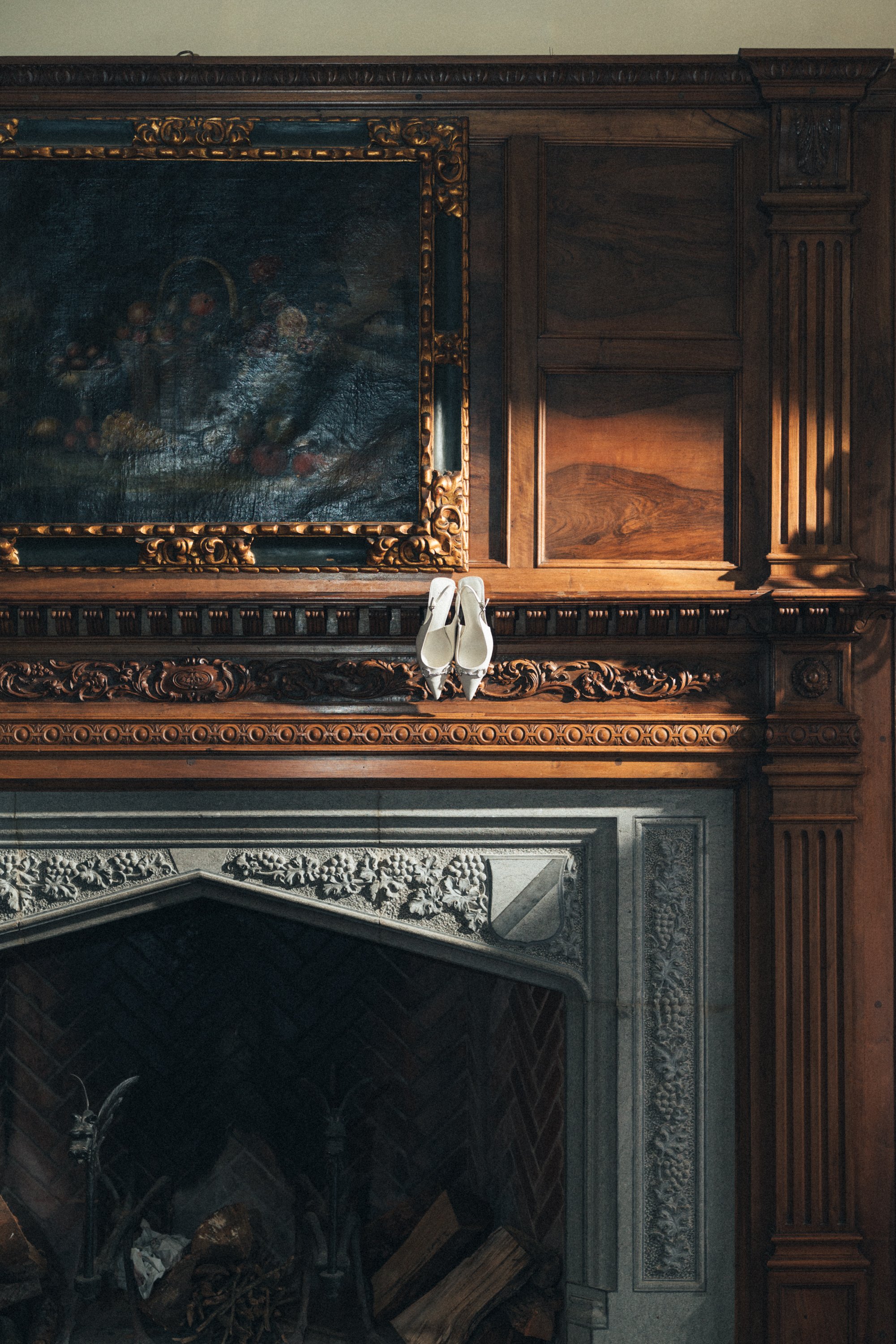 Ornate fireplace with a pair of white shoes on the mantel and a large framed painting above.