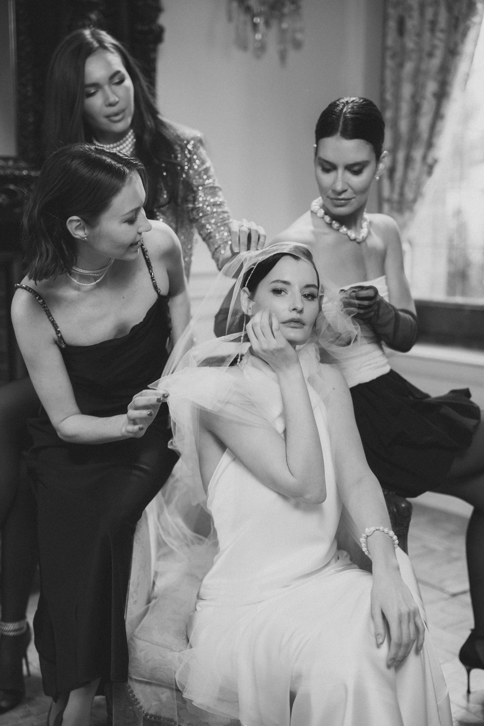 Black and white photo of a bride in a white dress sitting, surrounded by three women adjusting her veil and dress. The setting appears elegant, with luxurious decor.