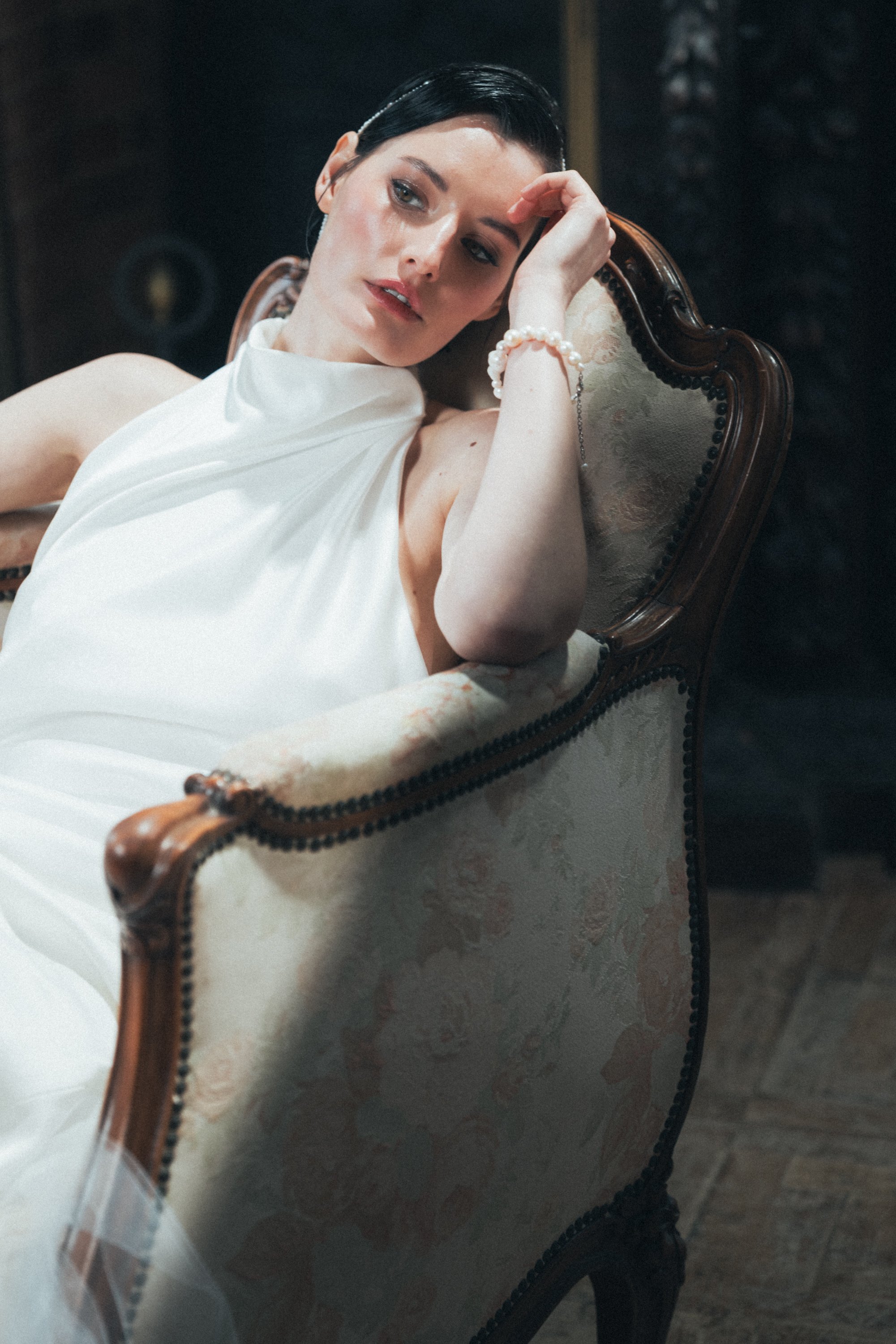 Woman in white dress sitting on ornate chair, resting head on hand.
