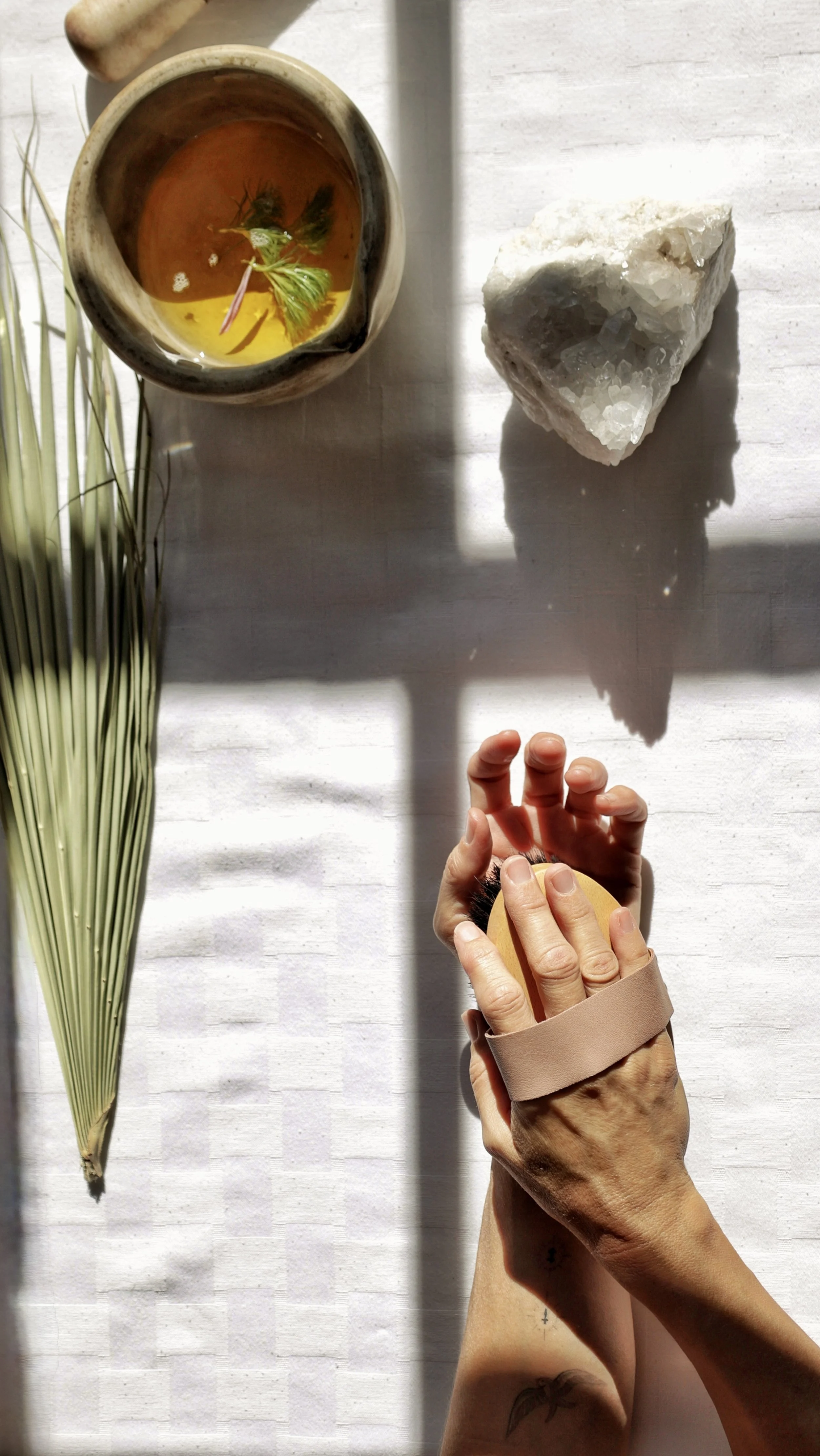 A hand holding a wooden brush over a white table with a container of herbal oil, a large white mineral rock, and green plant leaves.