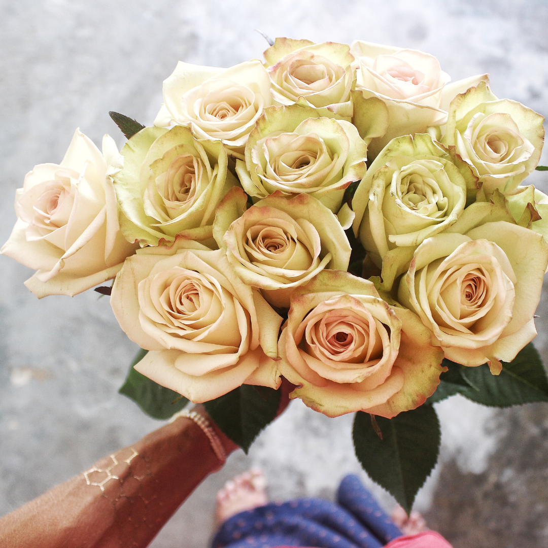 A bouquet of cream and blush pink roses with green leaves, held by a person wearing a brown sleeve and pink sandals on a gray concrete background.