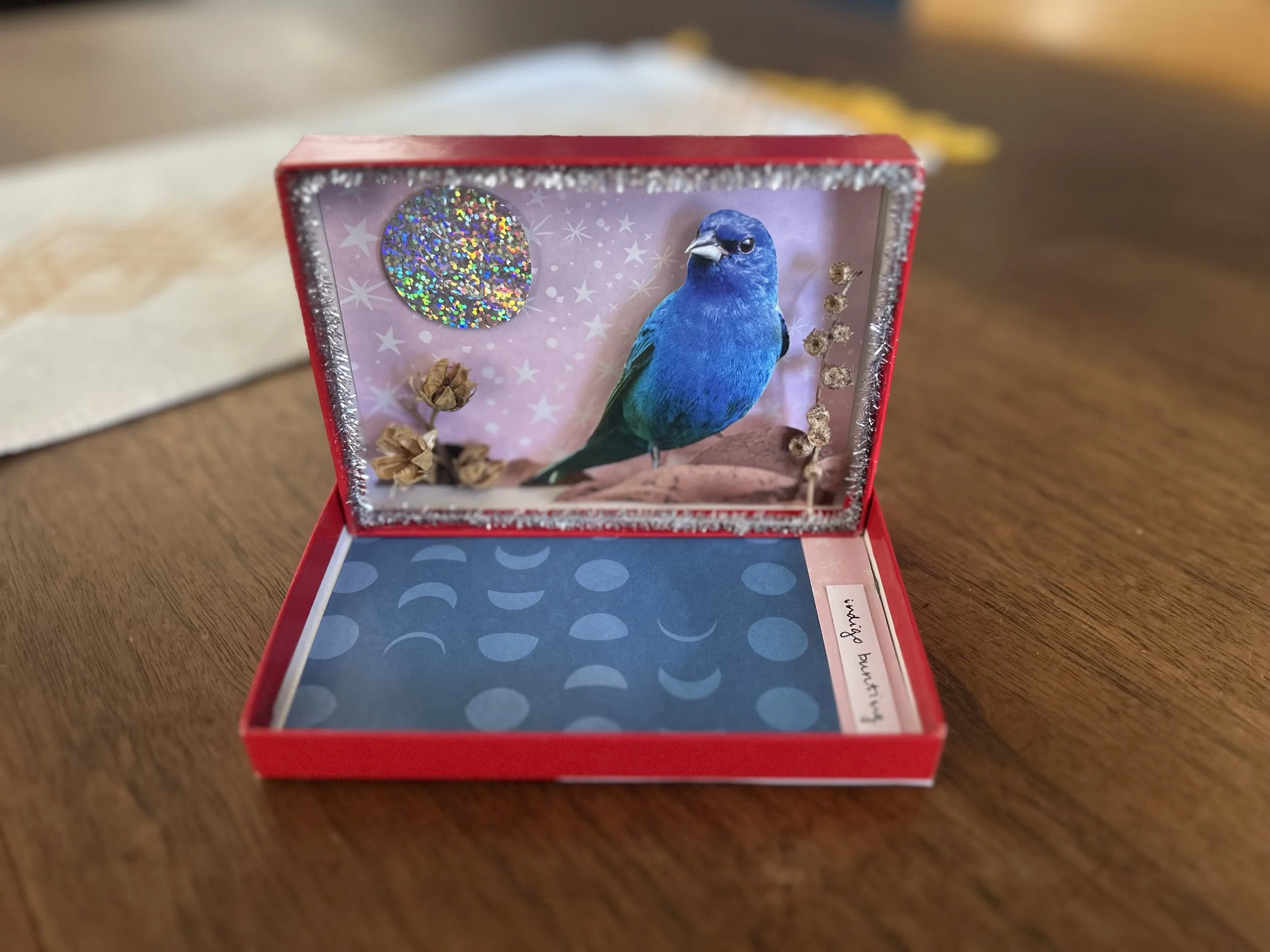 Open pink box with a 3D picture of a blue bird inside, decorated with glittery stars and dried flowers, on a wooden surface.