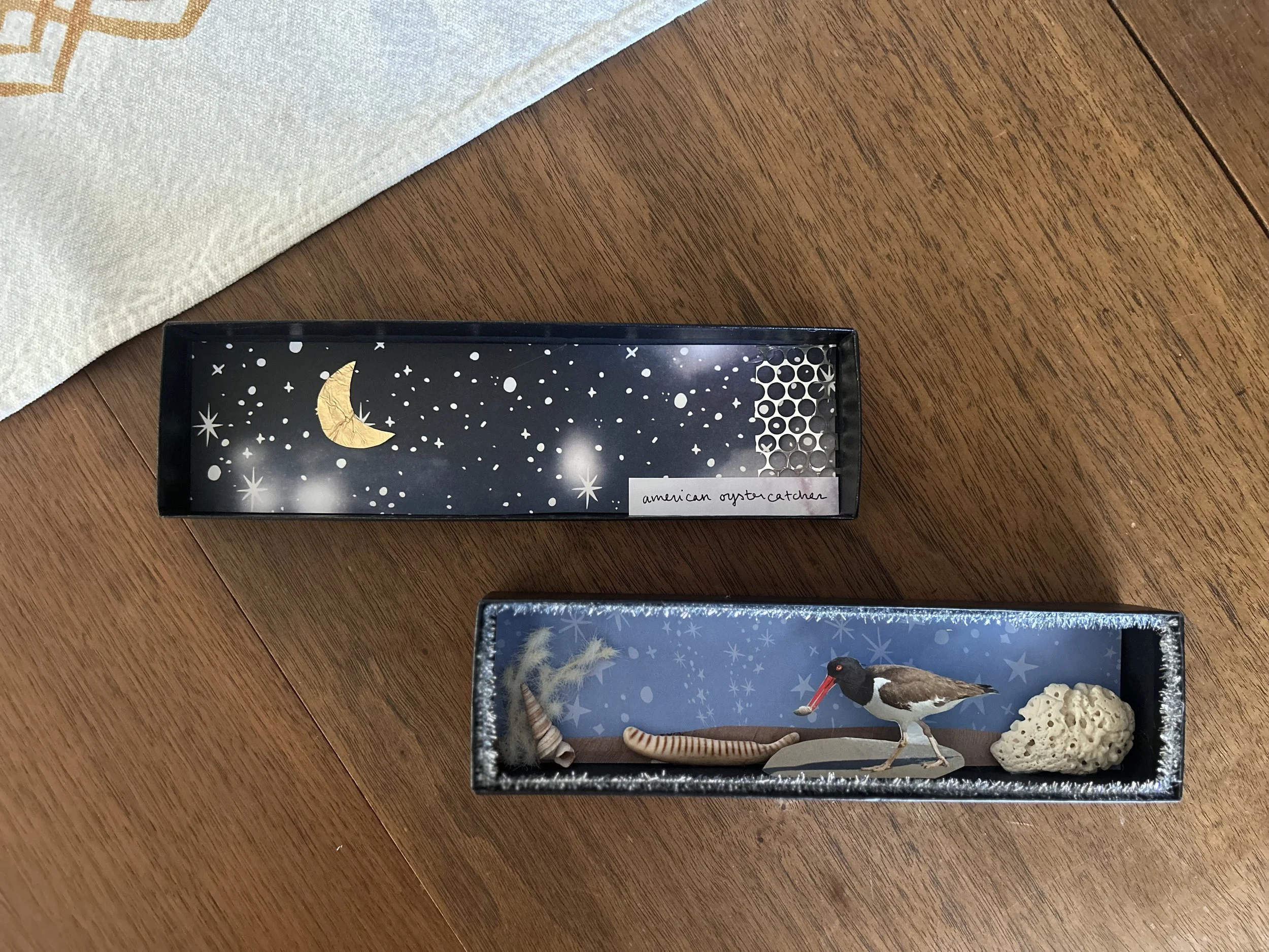 Two decorated shoeboxes with celestial and nature themes, containing small objects and figurines, placed on a wooden surface.
