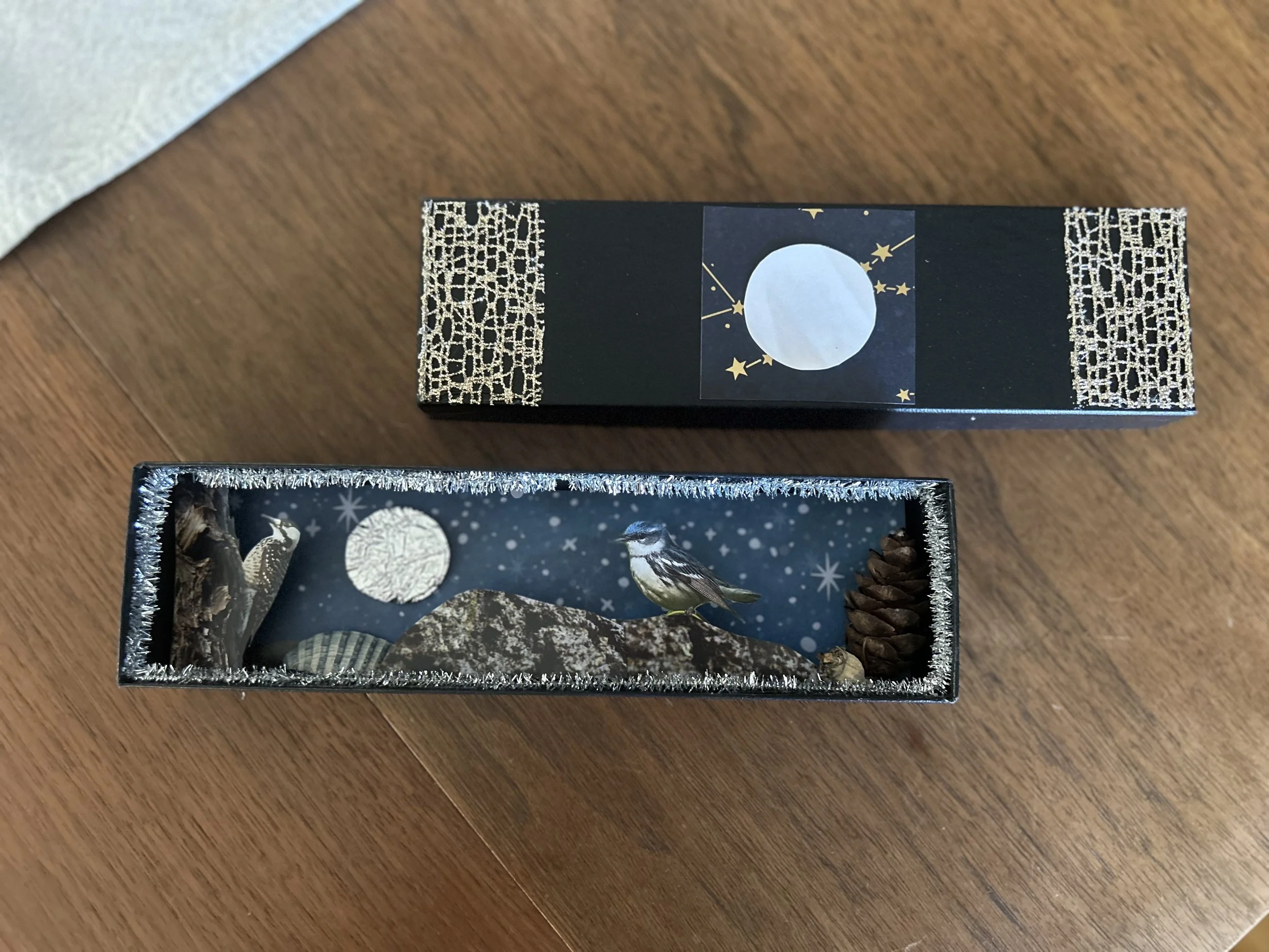 A miniature scene inside a decorated box featuring a bird perched on a faux rock, a pinecone, and a background with a moon and snowflakes, with the box lid placed above.