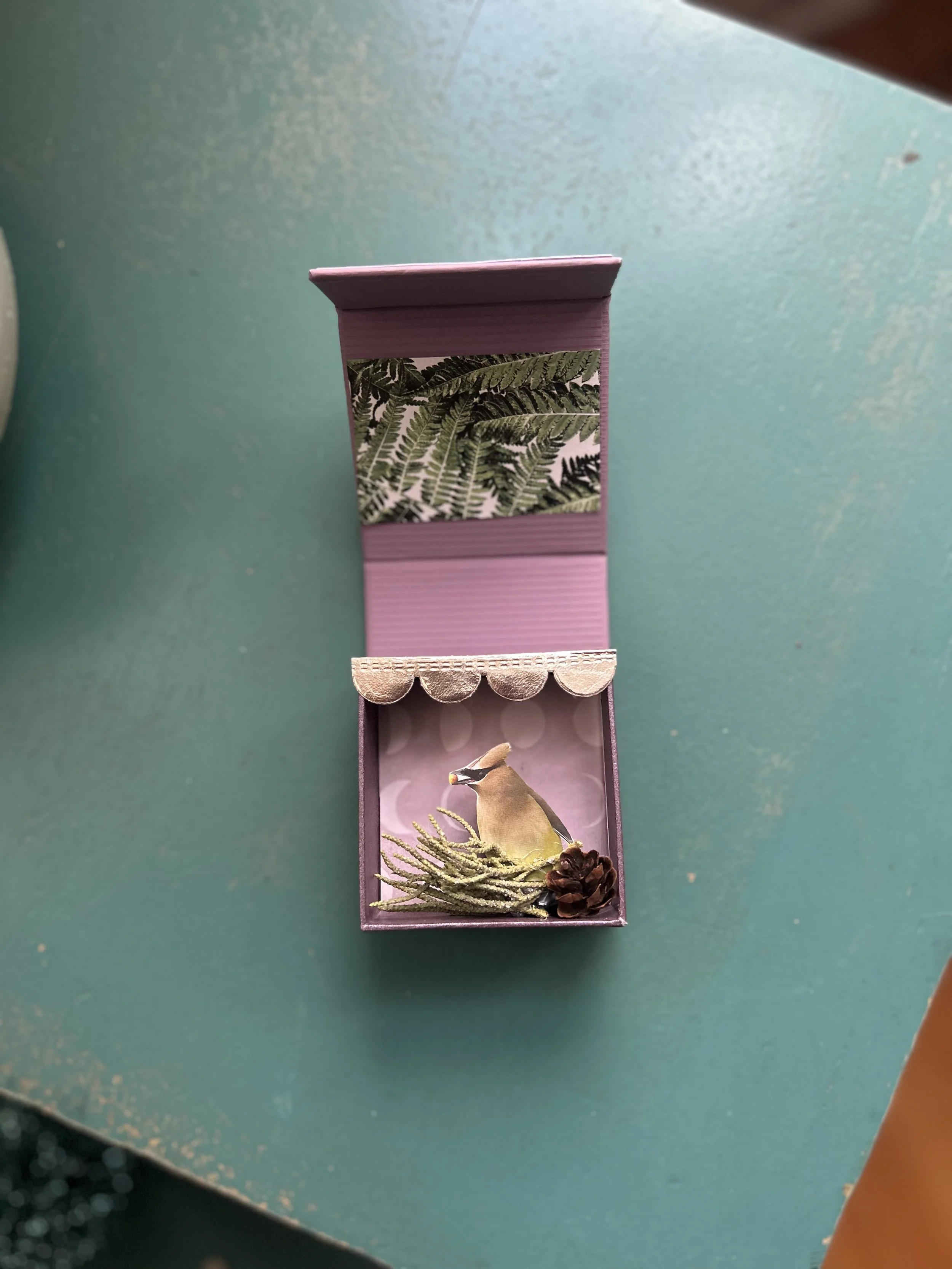A small, purple gift box with a scenic interior featuring a wooden bird figurine, a pine cone, and a sprig of greenery, with a leafy decorative cover on the lid.