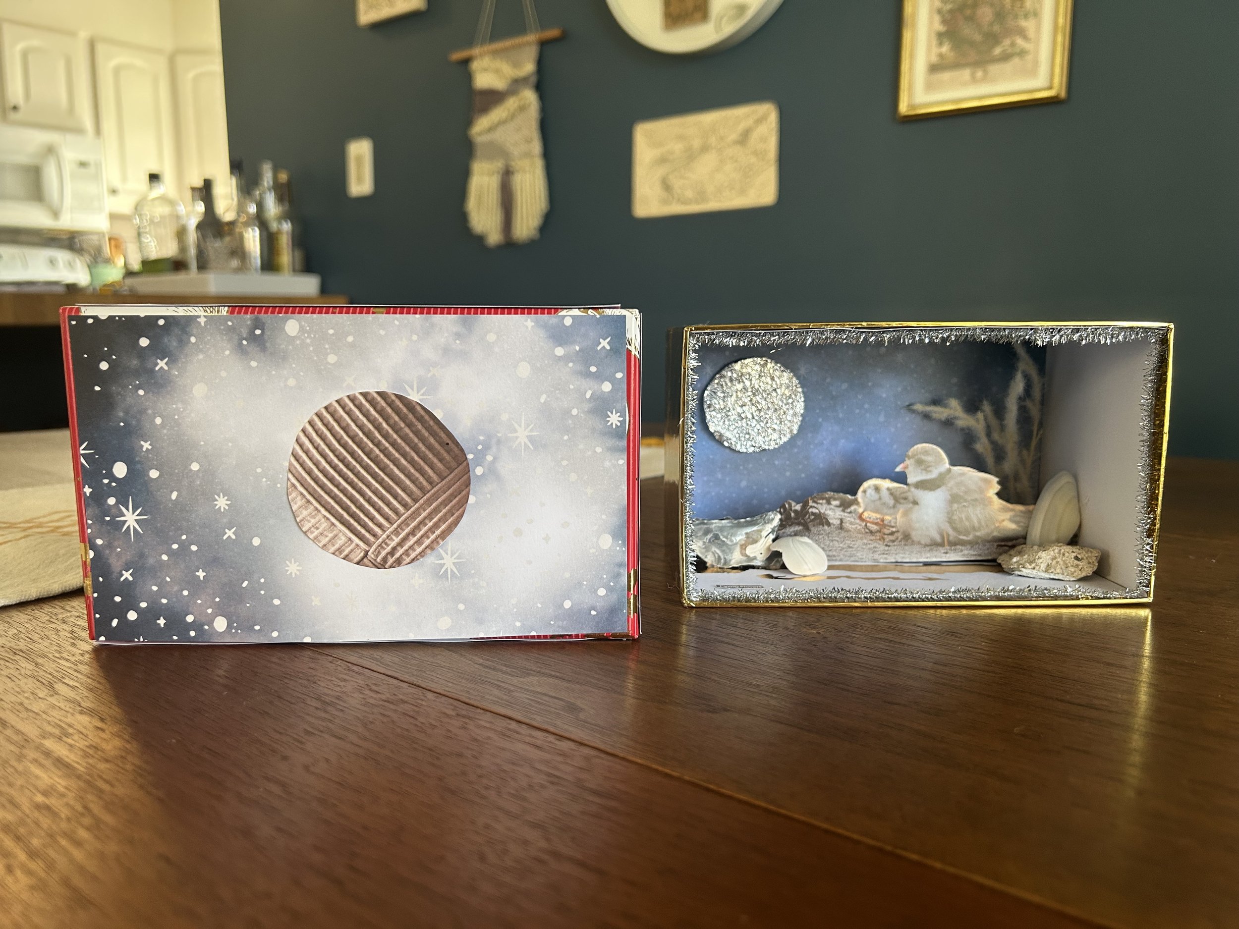 A miniature nativity scene inside a box with a snowy night sky background, featuring a moon, two baby ducks, shells, and rocks. Next to it is a cardboard box with a circular window and snowflake designs.