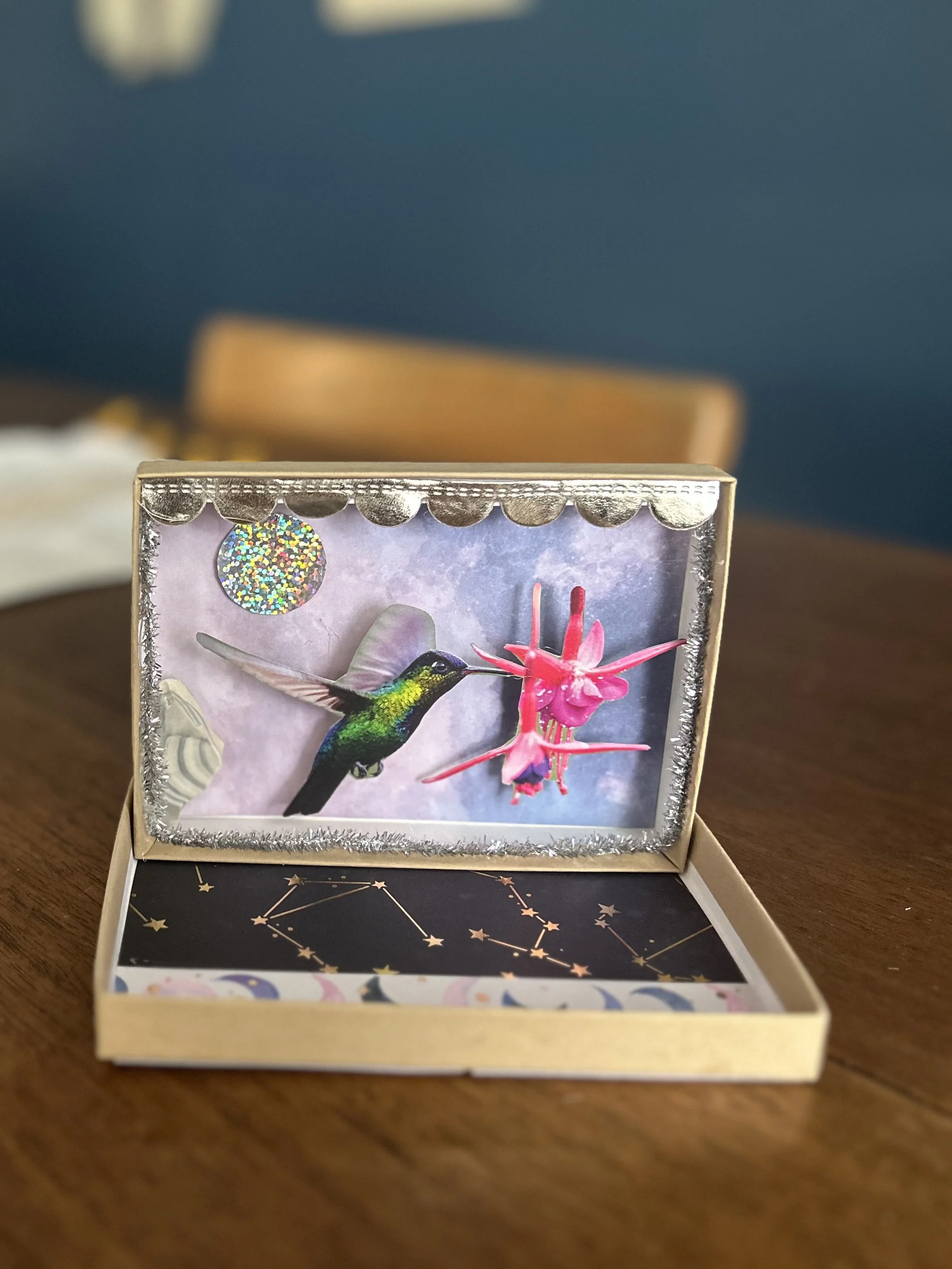 A jewelry box with a picture of a hummingbird drinking nectar from a pink flower, decorated with silver tinsel and a holographic sticker, placed on a wooden table with a star constellation pattern inside the lid.
