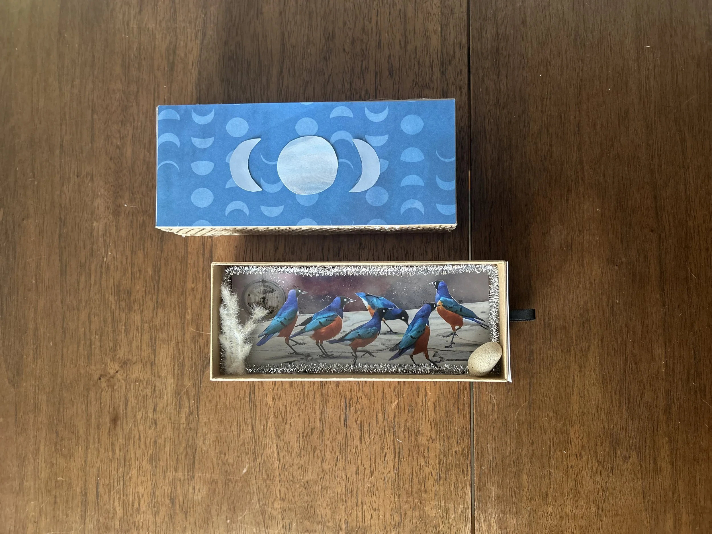 A decorative box with a blue lid featuring moon and star patterns, and an interior display of a small framed picture of five colorful blue and orange birds, decorated with silver tinsel, a small rock, and fluffy white material.