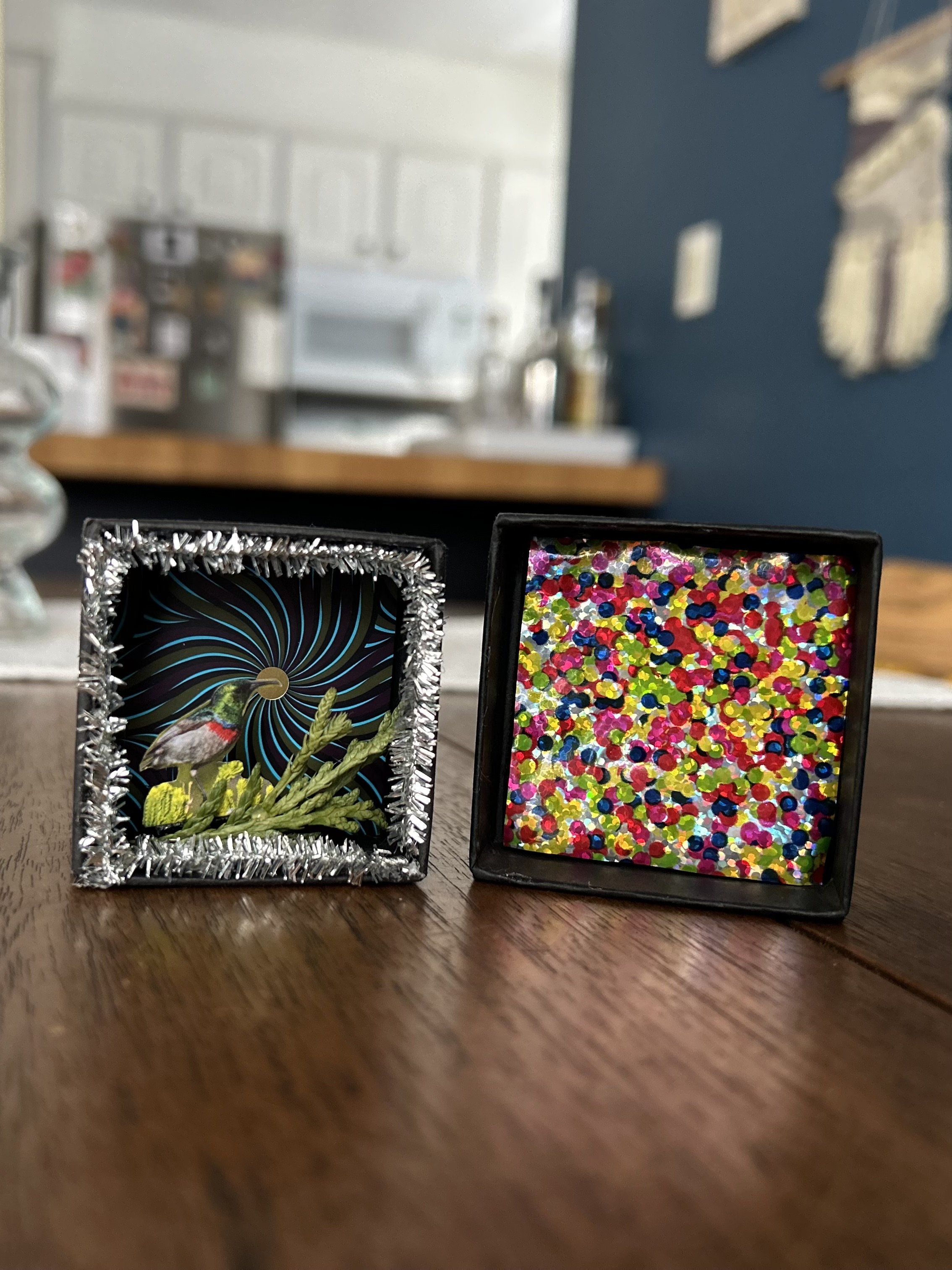 Two small decorative boxes, one with a holographic design and a bird image, the other filled with colorful confetti, placed on a wooden surface in a kitchen.