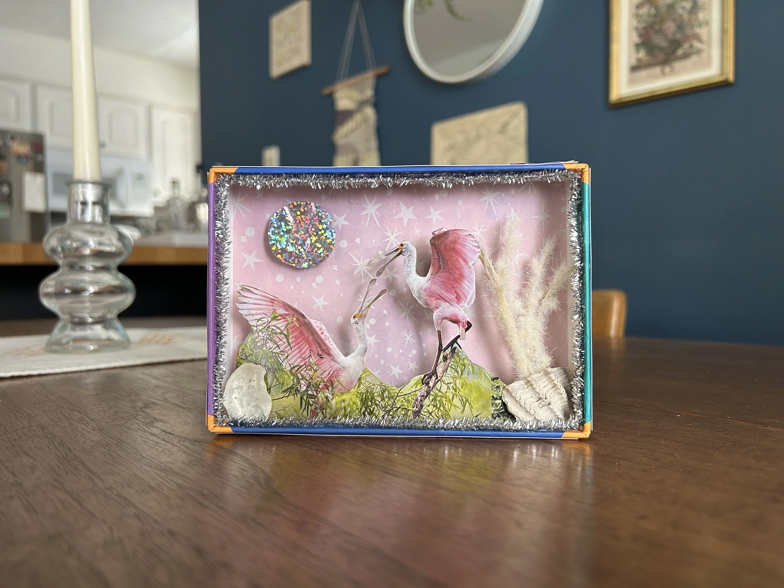 Decorative diorama of pink and white herons, greenery, shells, and a glittery circular ornament inside a display box on a wooden table.