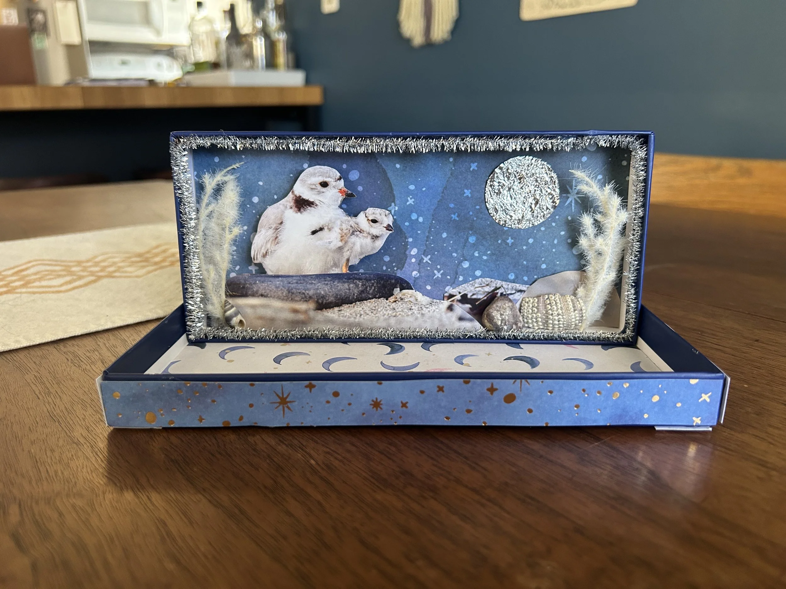 A decorative winter-themed card or artwork in a blue box with Christmas motifs and a snow scene, featuring a mother owl and owl chick, along with shells, artificial plants, and a moon in the background.