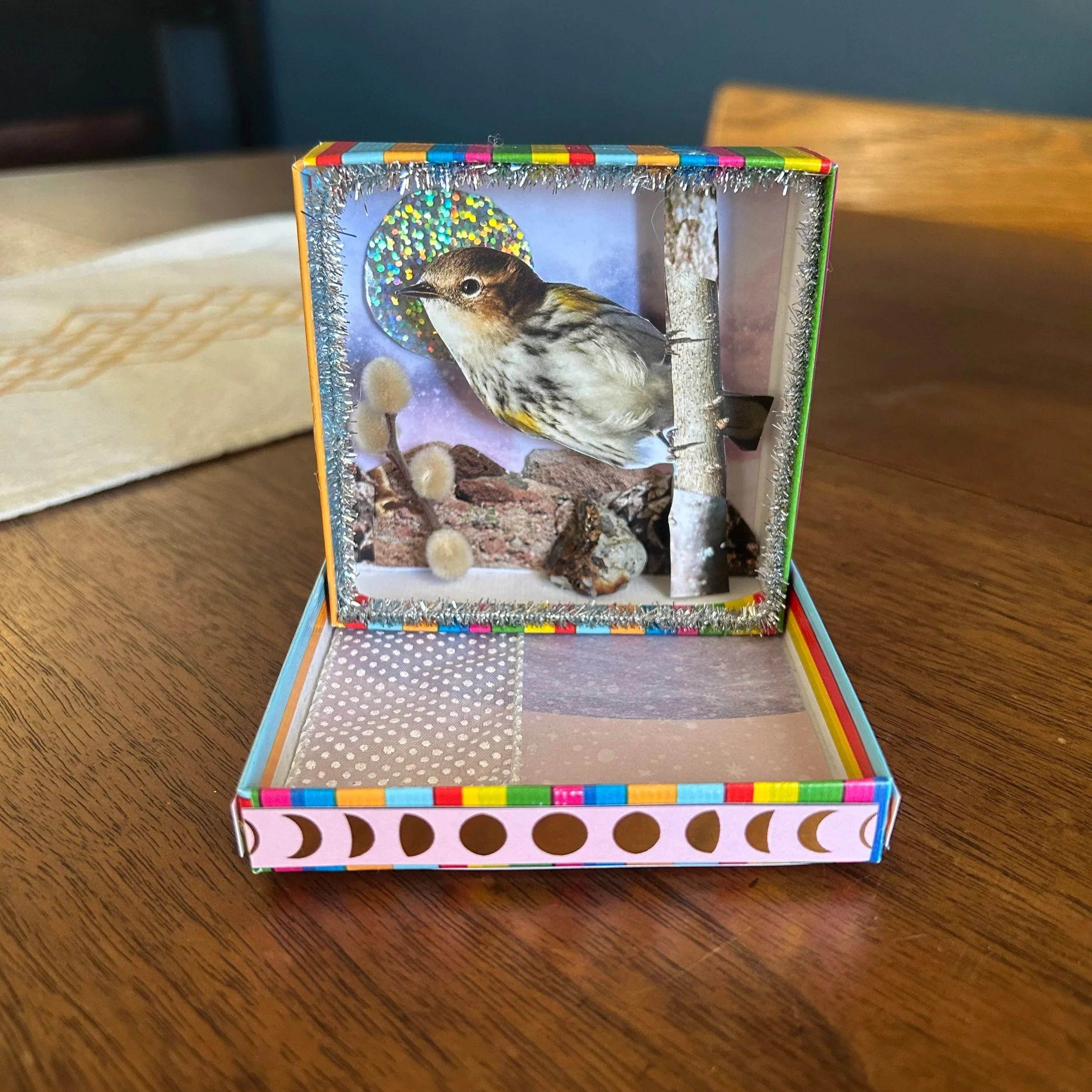 Decorative box with a cutout of a bird and background scene, featuring tinsel border and painted dots.