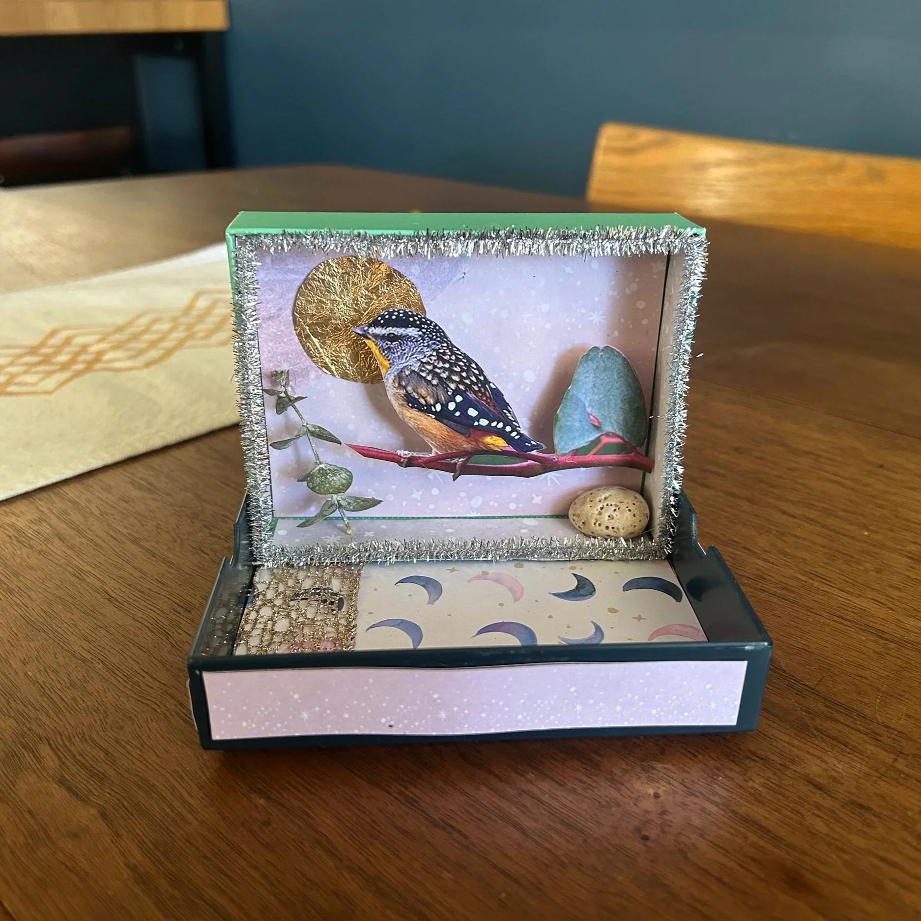 A decorative box with a bird illustration inside, featuring a bird perched on a branch against a festive background with snowflakes, gold accents, and moon patterns.