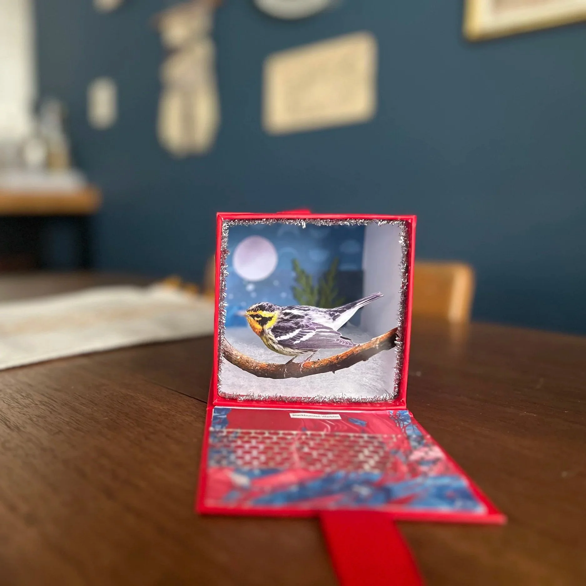 A small decorative box with a picture of a bird on a branch inside it, set on a wooden table. The box's interior is decorated with fake snow and a winter scene in the background.
