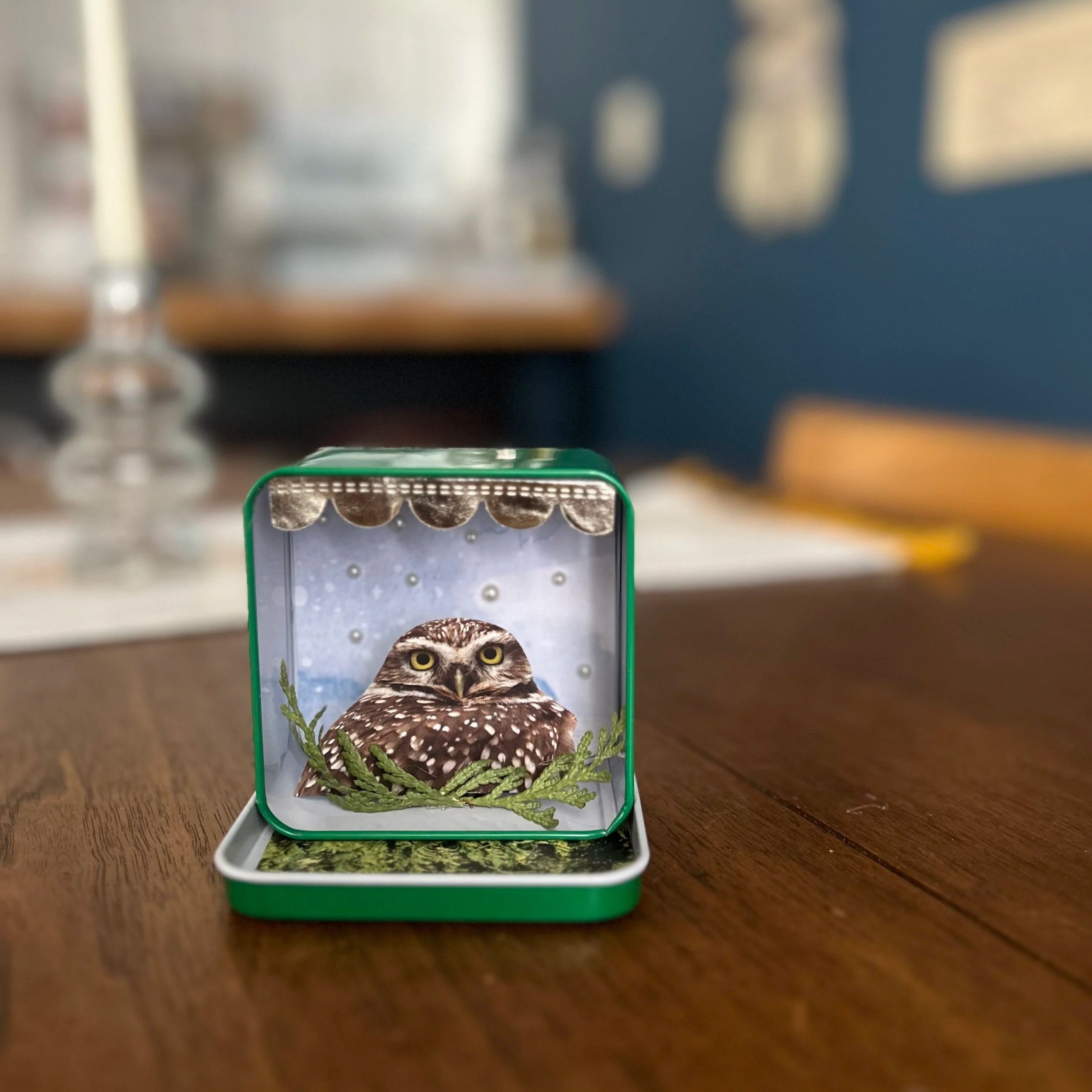 Decorative item featuring an owl with yellow eyes and brown feathers, surrounded by green foliage, inside an open metal container.