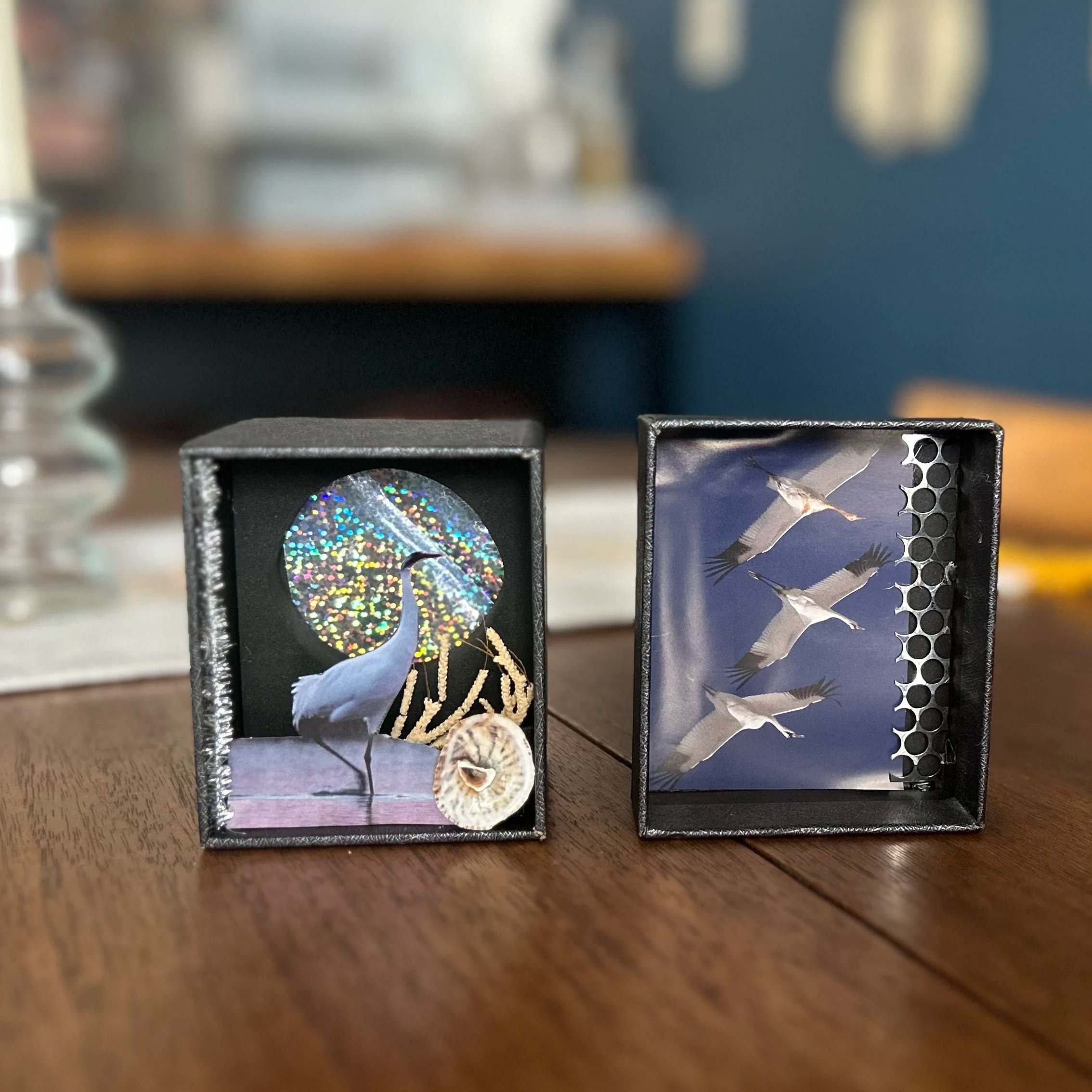 Two small boxes with fairy tale or fantasy-themed decorations on a wooden surface. The left box features a white heron, a holographic circle, a seashell, and golden string or decorations inside. The right box displays a photo of flying cranes against