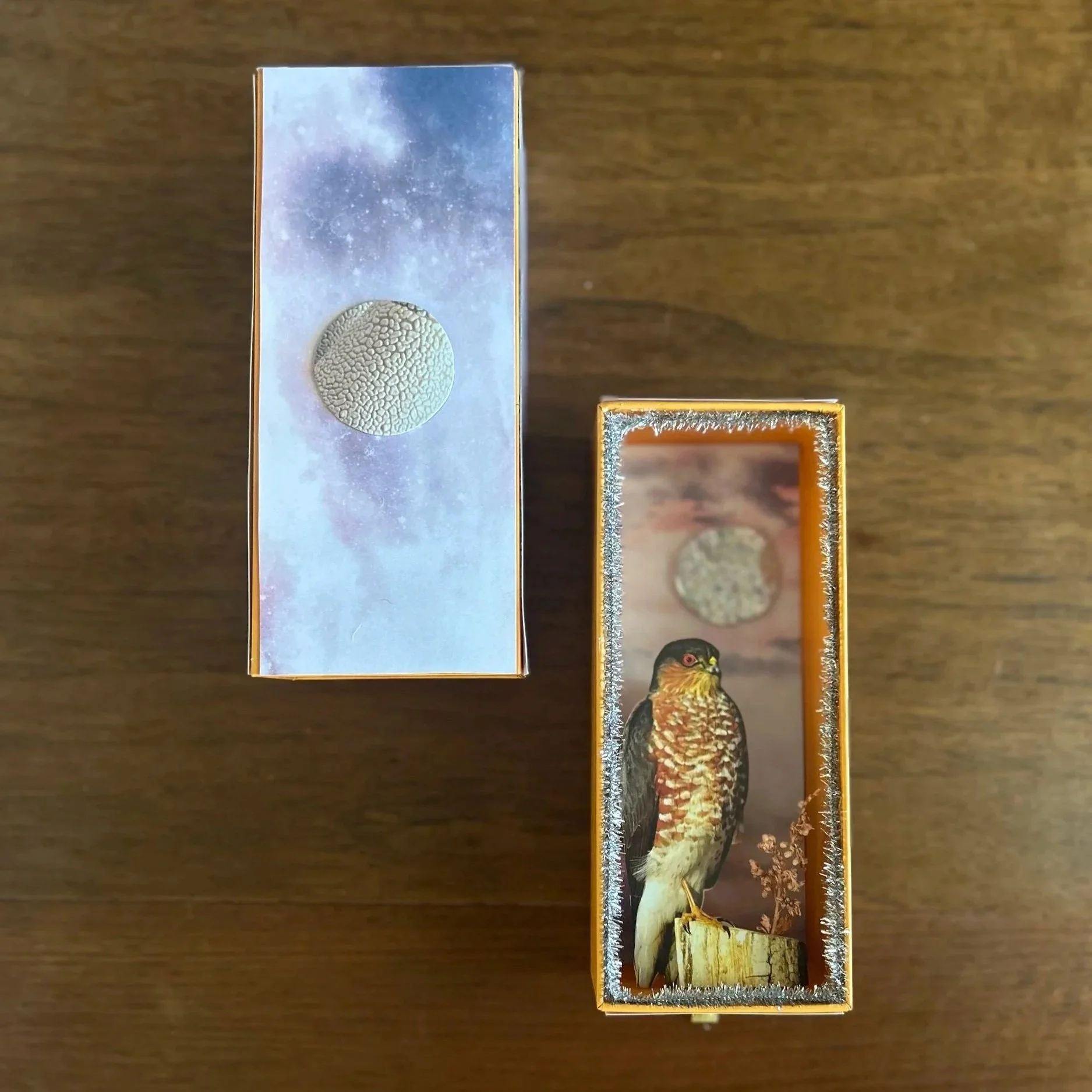 Two decorative frosted matchboxes on a wooden surface, one with a moon illustration and the other with a bird illustration.