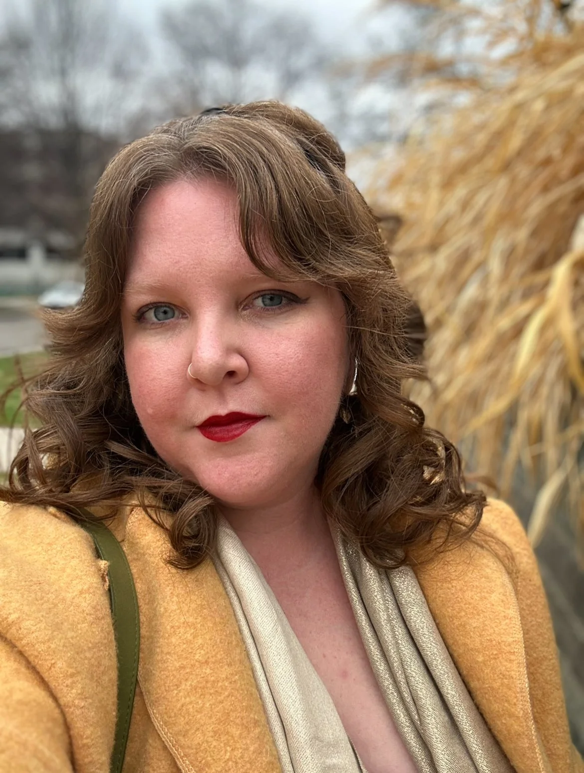 A woman with curly brown hair, red lipstick, and a nose ring taking a selfie outdoors in front of dried yellow plants, with a blurred background of trees and a cloudy sky.