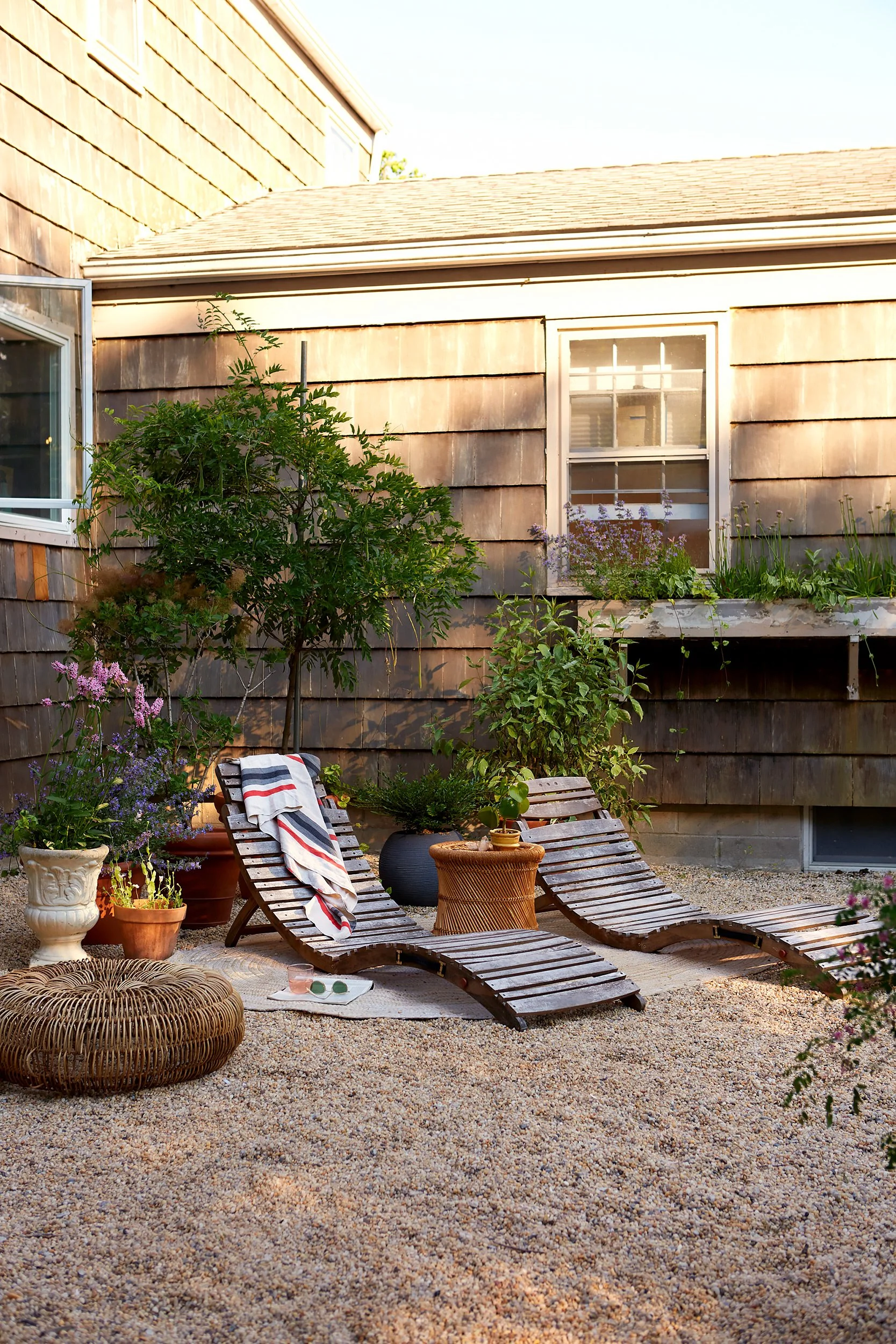 A cozy backyard scene with two wooden lounge chairs, potted plants, a wicker pouf, and a small table, set against a rustic wooden house exterior with a window and greenery.