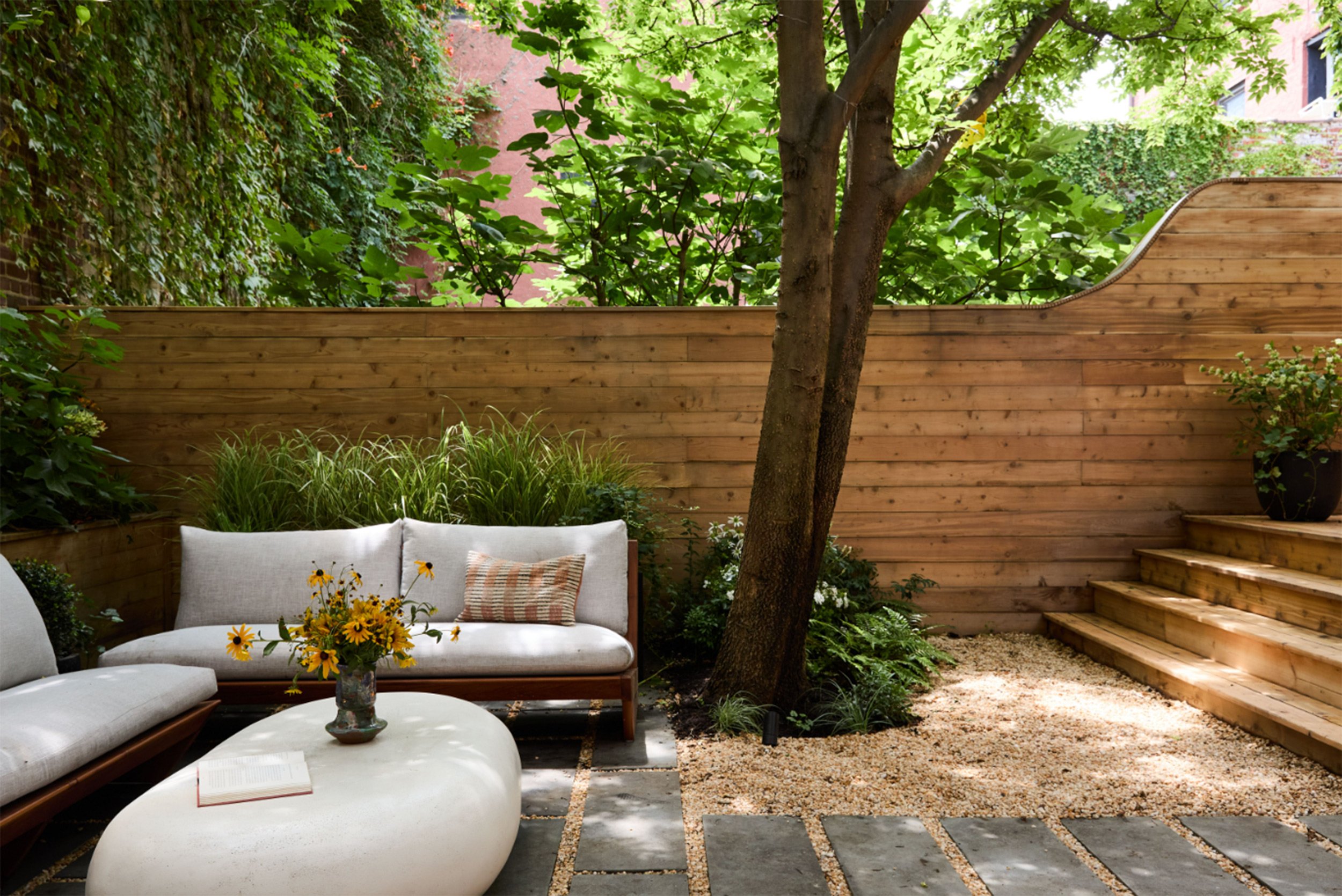 A cozy outdoor patio with a wooden fence, green plants, a tree, outdoor seating with cushions, a coffee table with a vase of yellow flowers, and steps leading up to an elevated garden area.