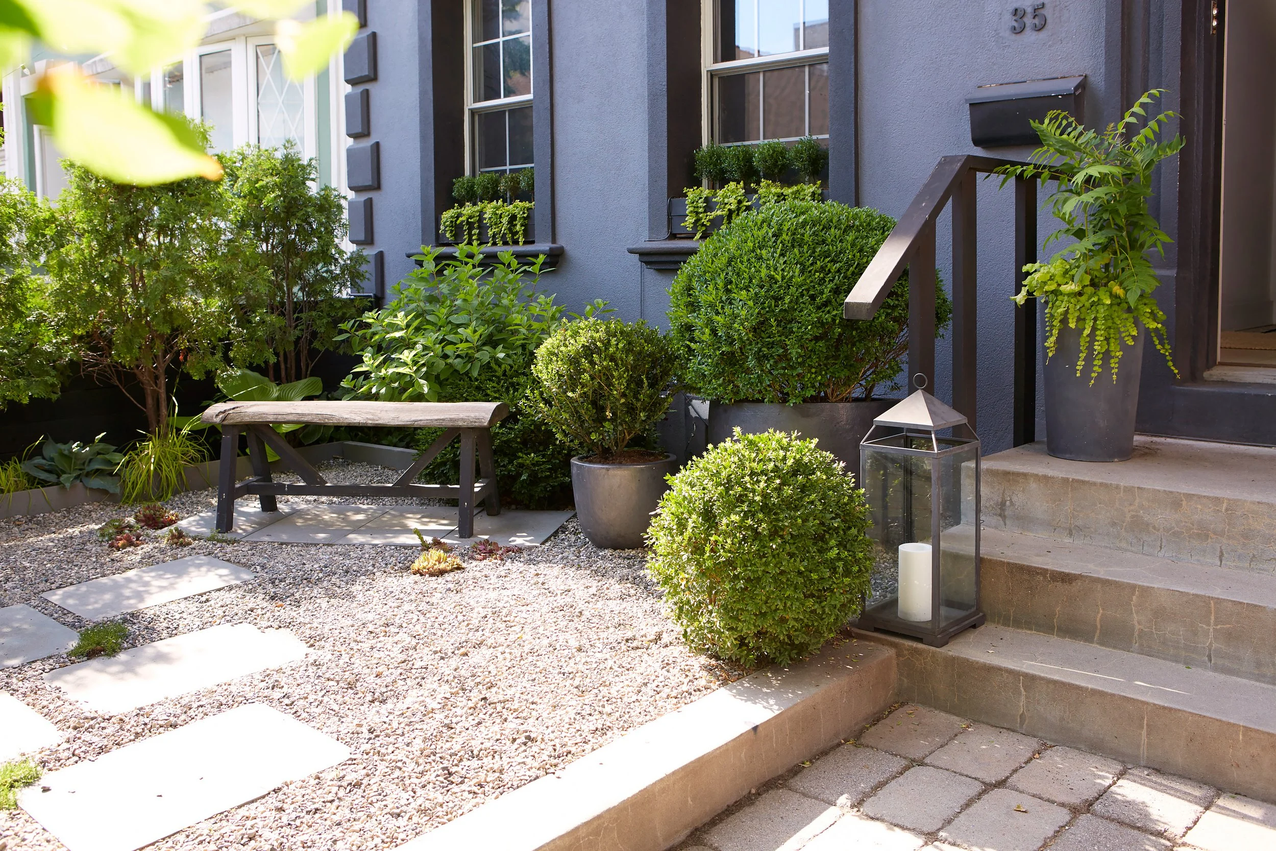Front porch of a house with a stone pathway, green plants in pots, a lantern with a candle, and stairs leading to the door.